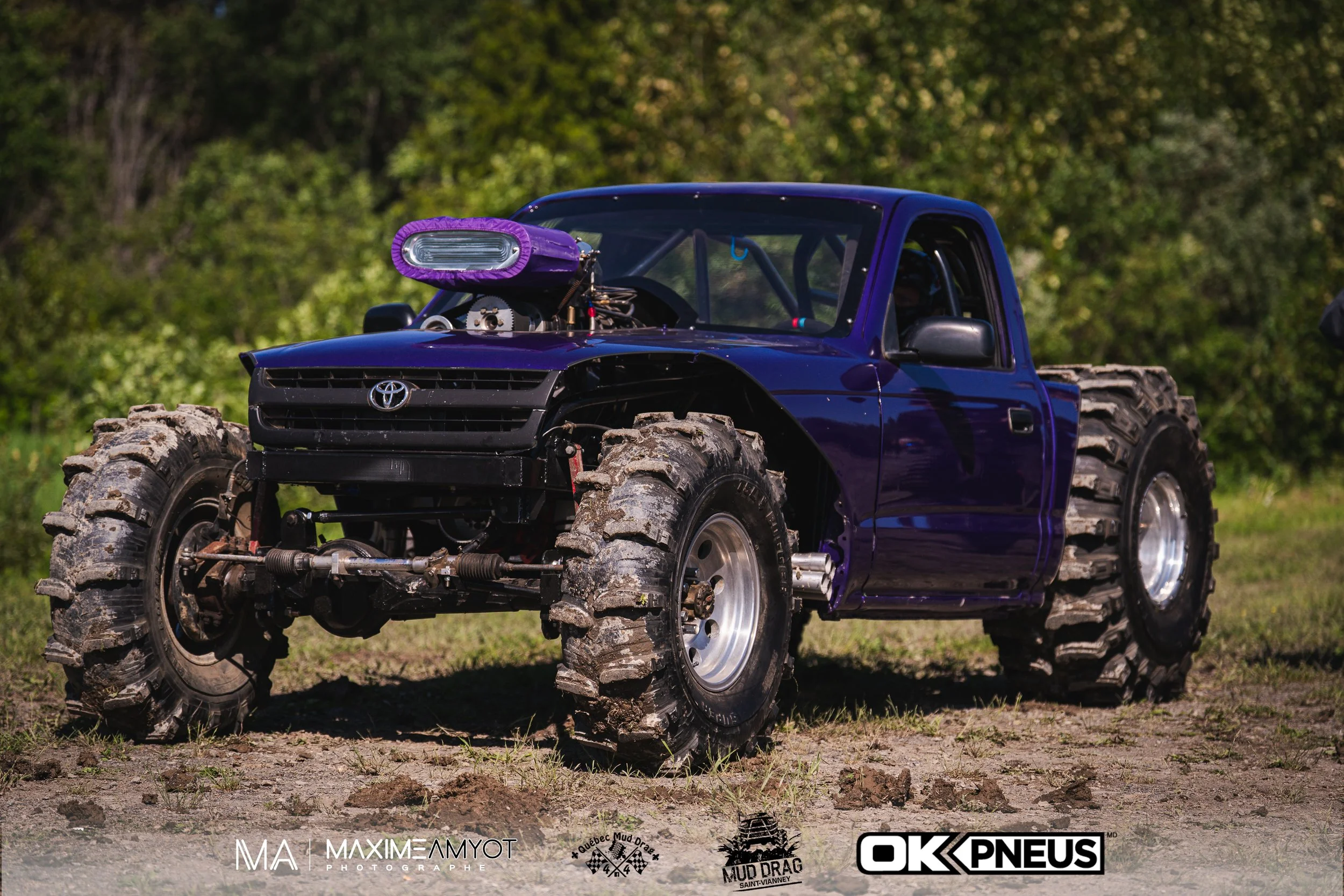 Un truck modifié pour le mud drag avec de grosses roues tout-terrain, un moteur exposé avec un cache en tissu violet, et une carrosserie bleu foncé, sur un terrain poussiéreux avec de la végétation en arrière-plan.