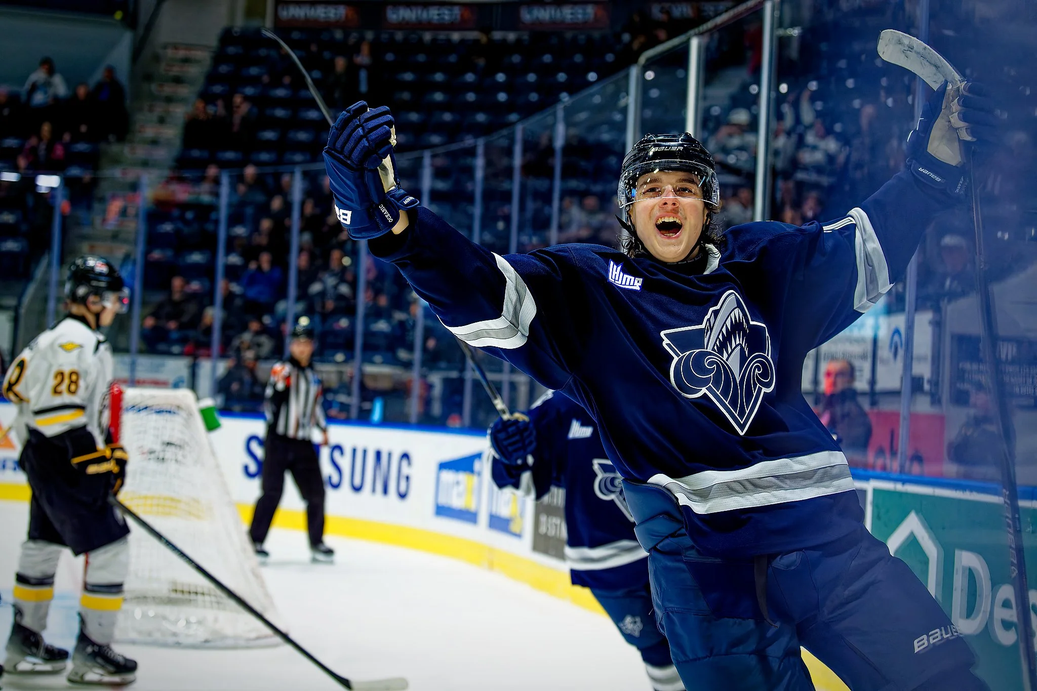 Ice hockey player in navy blue jersey celebrating with arms raised on ice rink, with other players and audience in the background.