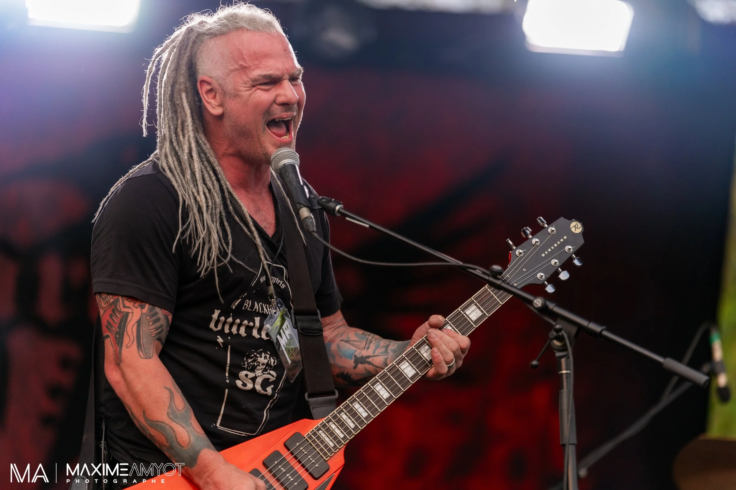 Un homme avec des dreadlocks grises chante en jouant de la guitare électrique sur scène.