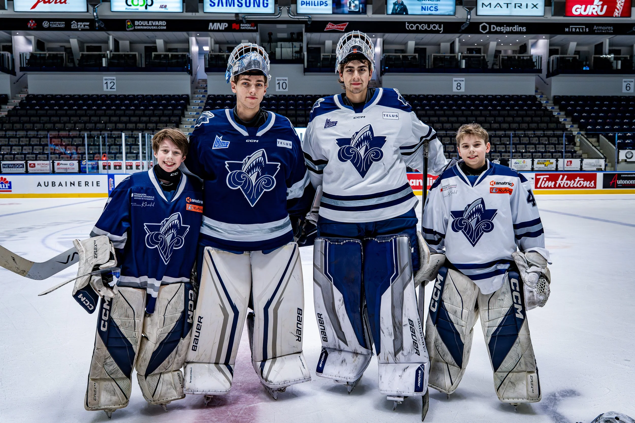 Cinq jeunes hockeyeurs posant sur la glace dans une arène, portant des équipements de hockey et des maillots avec un logo de fendrière. Deux gardiens de but au centre, deux jeunes joueurs sur côtés, un jeune joueur en uniforme et arme de hockey.