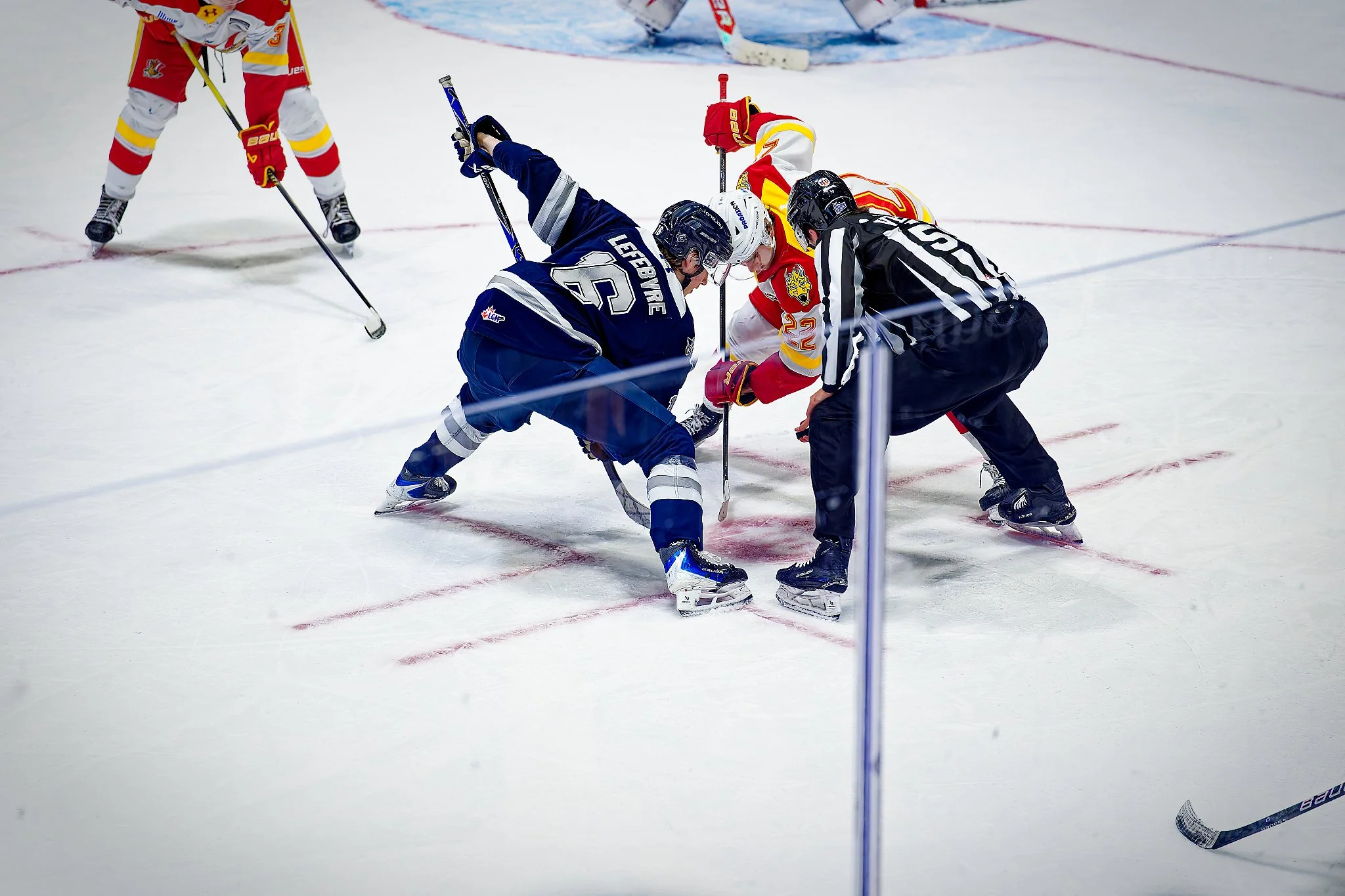 Two hockey players, one in a dark blue jersey and the other in a white jersey with red and yellow accents, are fighting for the puck at the faceoff circle while a referee watches by their side; a third player in red and white is skating nearby on the