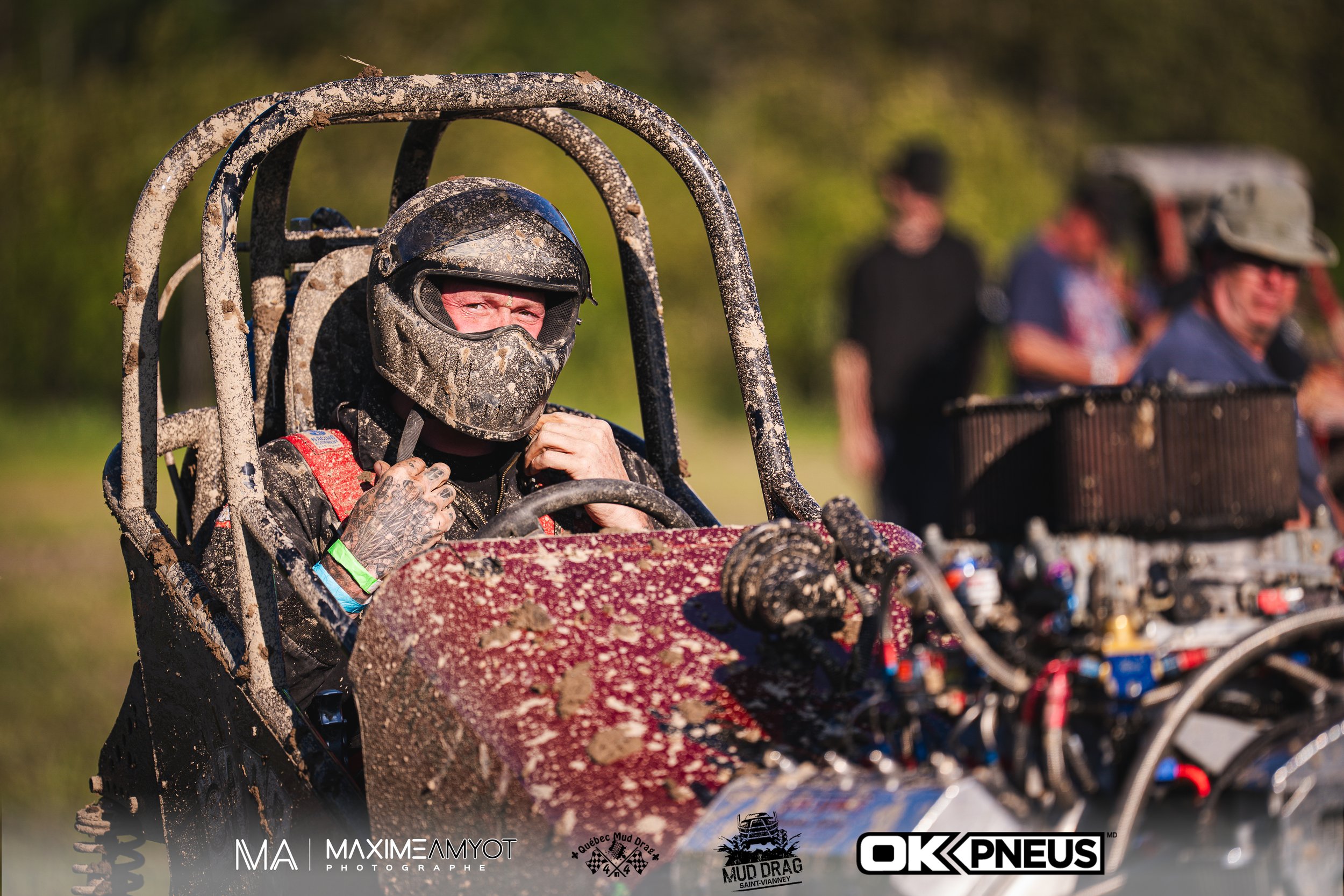 Pilote d'un buggy tout-terrain couvert de boue, portant un casque et une combinaison de protection, assis dans le véhicule avec d'autres personnes en arrière-plan.