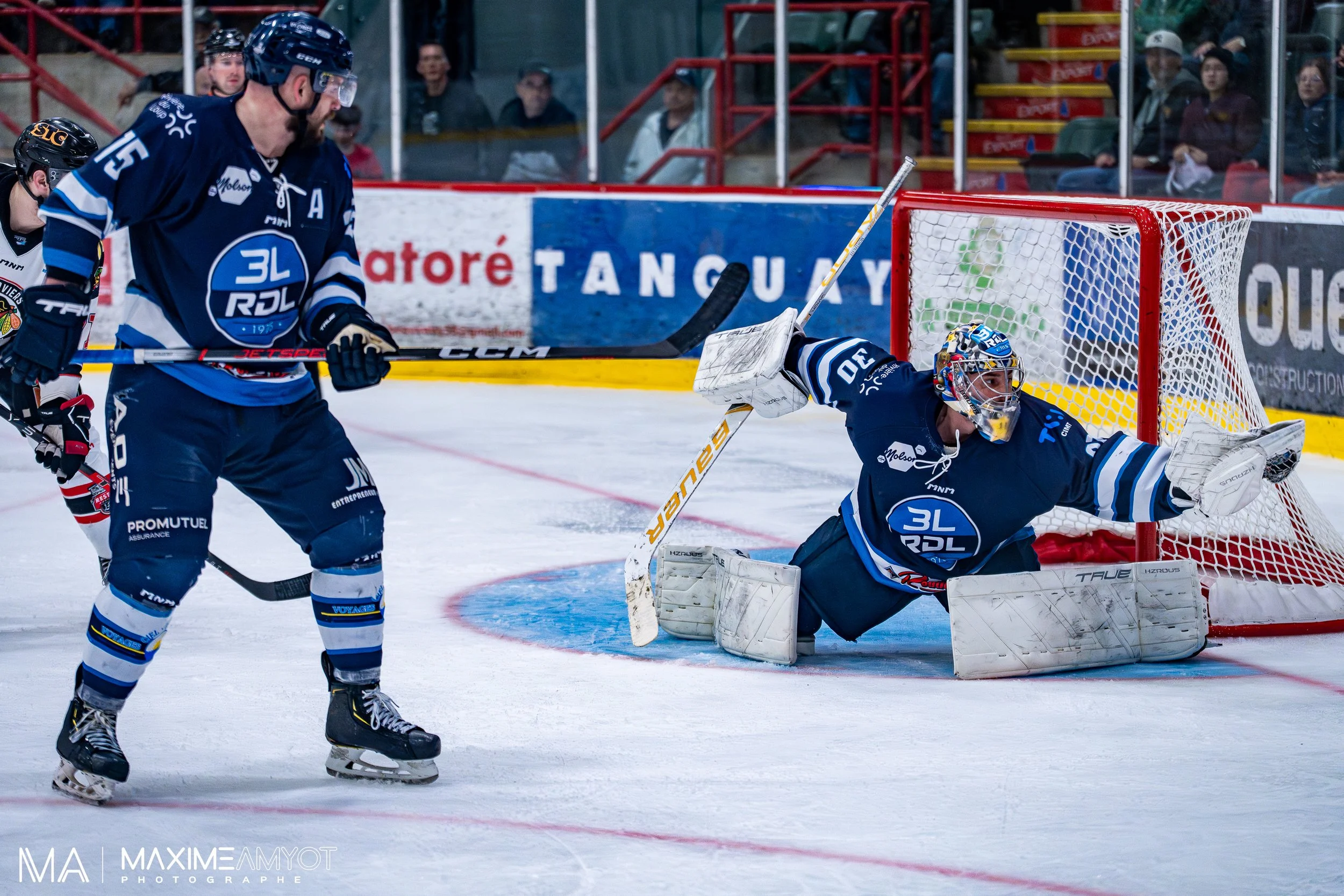 Une scène de hockey sur glace montrant un gardien en pleine action face au but, avec plusieurs joueurs autour, dans une patinoire. Spectateurs regardent derrière la vitre.