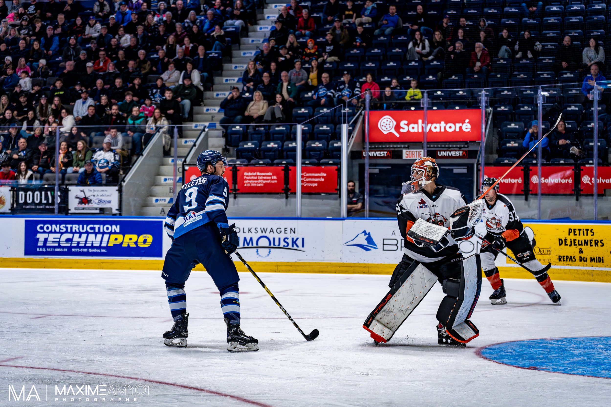 Match de hockey sur glace entre deux équipes, avec des joueurs en action sur la glace et des spectateurs dans les gradins.