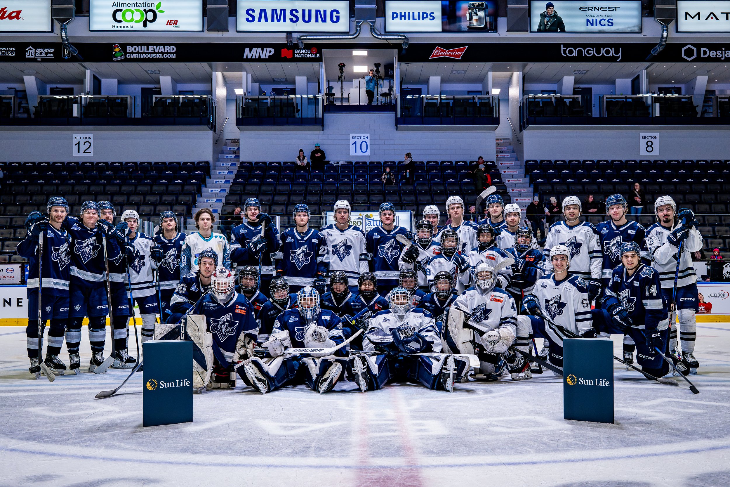 Équipe de hockey sur glace posant sur la glace dans un aréna, visages sérieux, tenues bleues et blanches, avec spectateurs en arrière-plan.