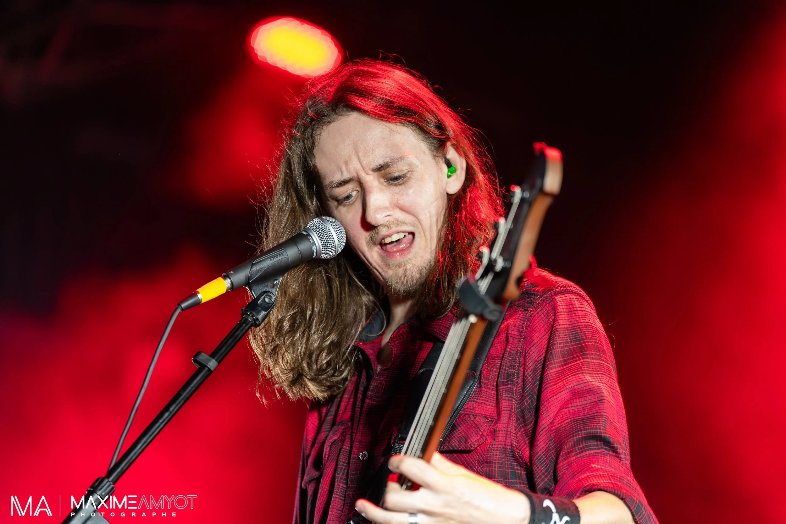 Musicien avec une guitare électrique, portant une chemise à carreaux rouges, chantant et jouant lors d'un concert, sous un éclairage rouge intense.