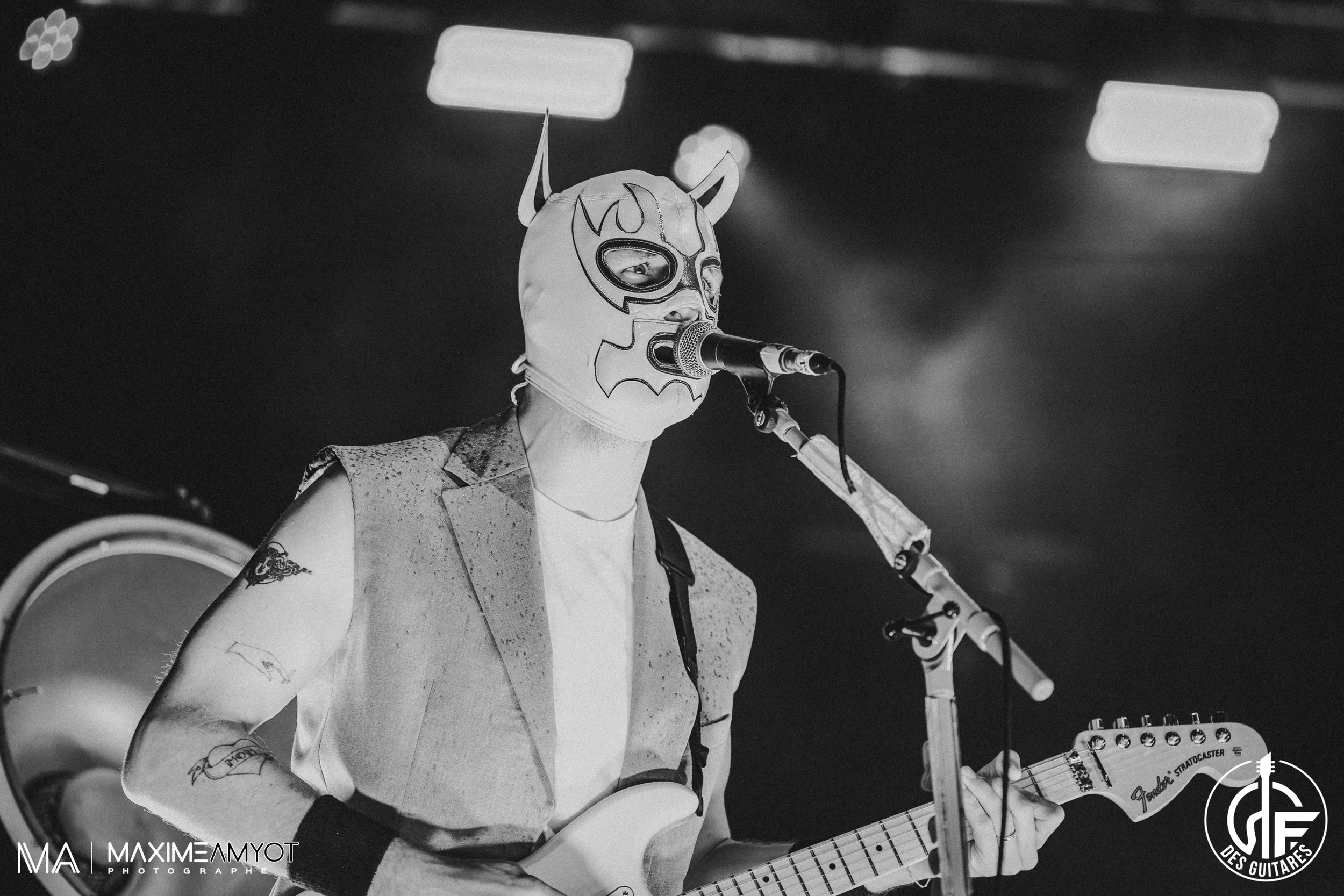 Musicien portant un masque de luchador jouant de la guitare électrique sur scène, noir et blanc.