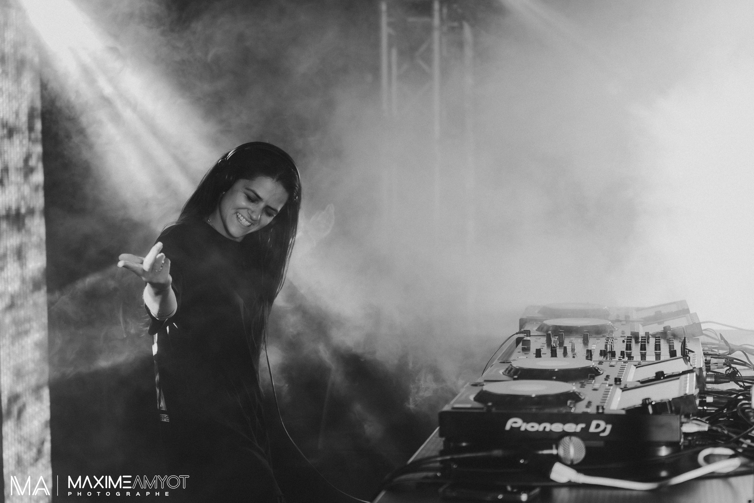 Une femme DJ souriante et jeune, portant un casque audio, joue avec la main près de plateaux de DJ, dans un environnement de scène avec fumée et éclairage.
