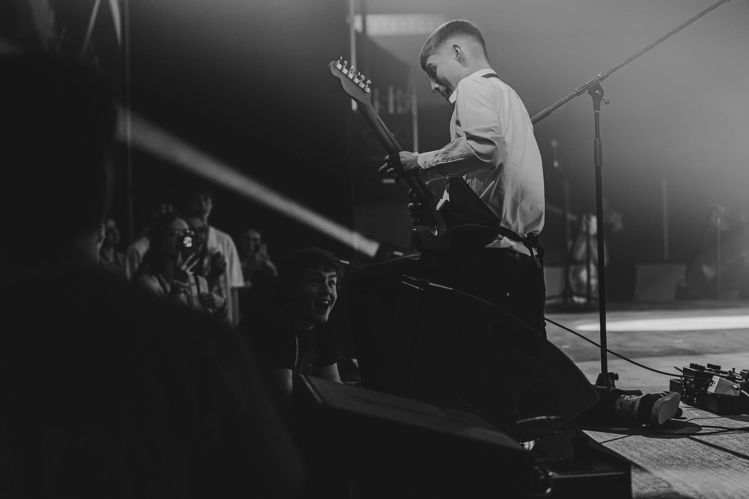 Musicien jouant de la guitare électrique sur scène lors d'un concert, photo en noir et blanc, spectateurs regardant et applaudissant en arrière-plan.