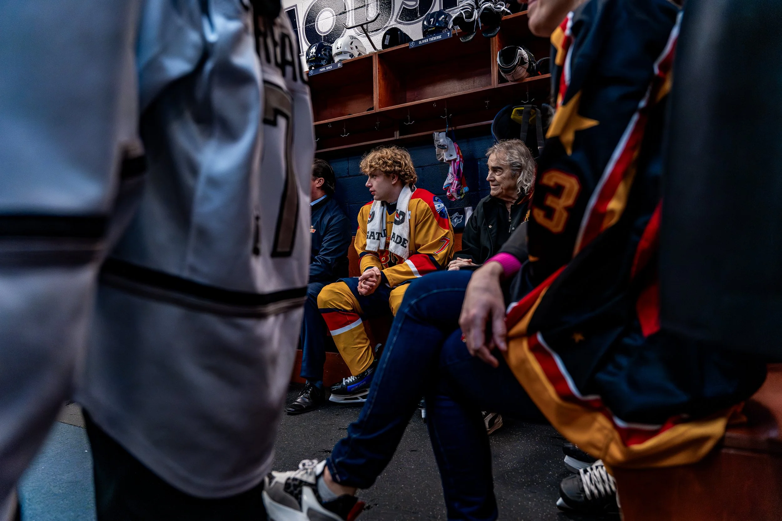 People sitting in a hockey locker room, some wearing hockey jerseys and jackets, with hockey helmets and gear on a shelf behind them, preparing for or reflecting after a game.
