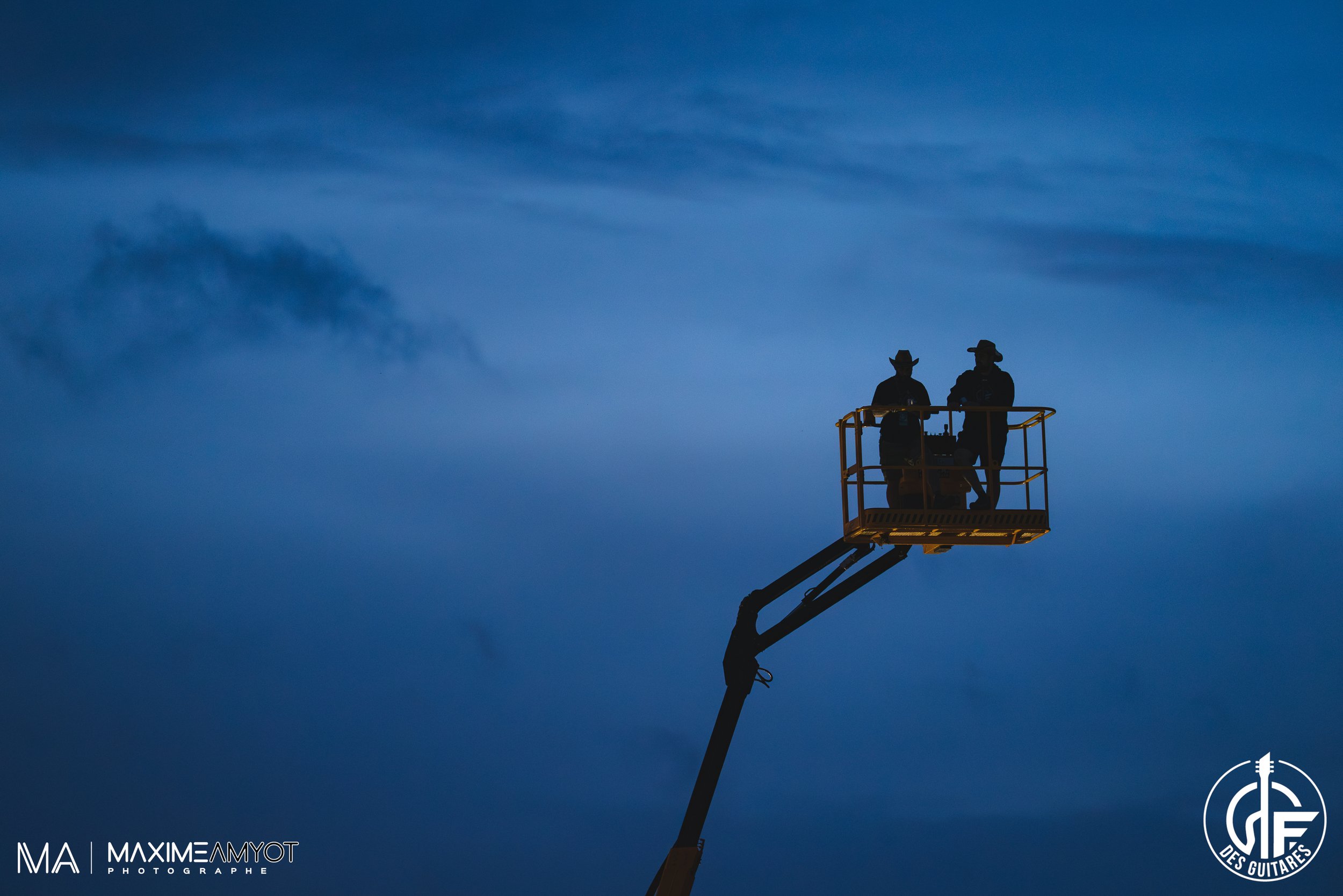 Deux personnes en silhouette sur une nacelle élévatrice, éclairant un ciel bleu nocturne, portant des chapeaux de cowboy.