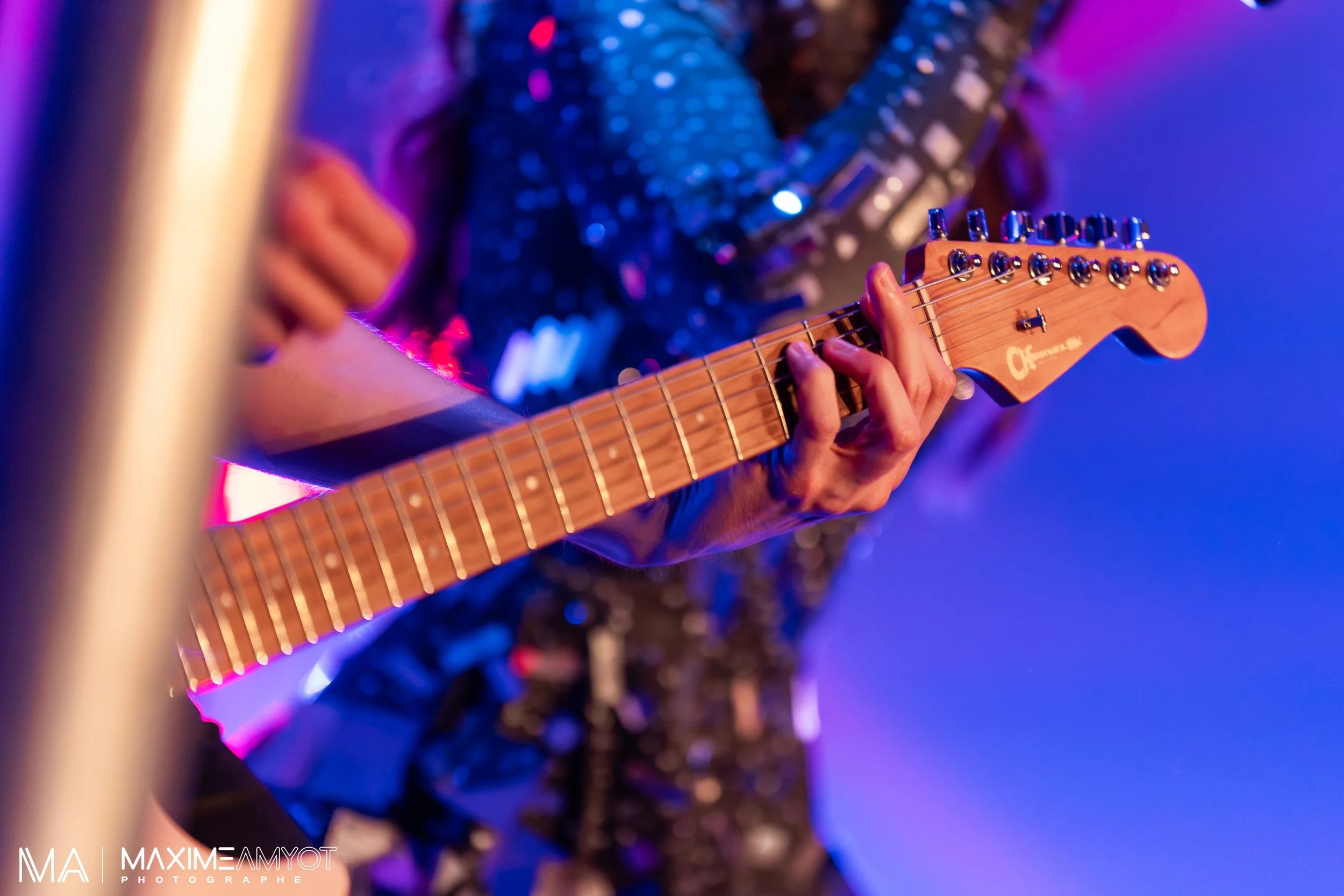Guitare électrique en bois naturel jouée par une personne lors d'une performance, avec un fond coloré en bleu et violet.