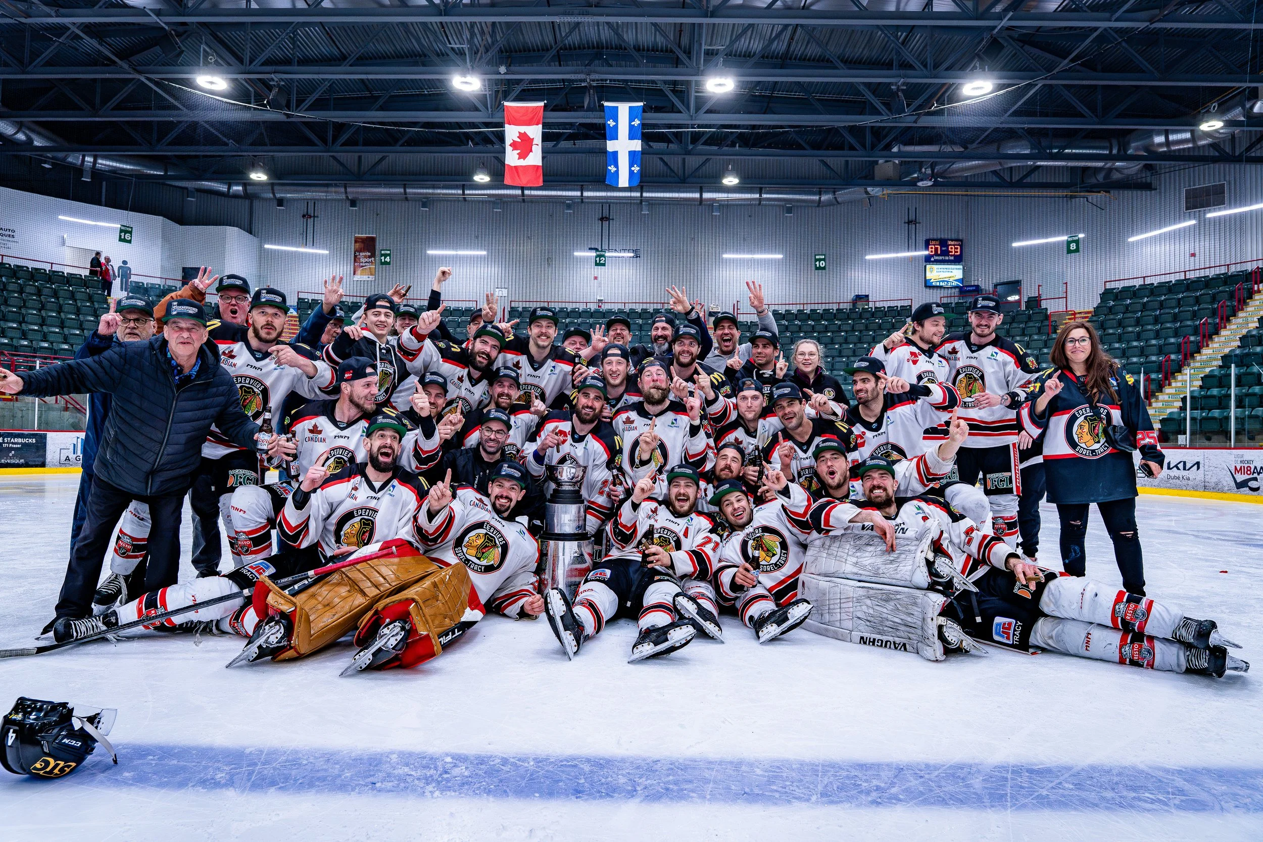 Une équipe de hockey sur glace en maillots célébrant leur victoire dans une patinoire, avec un trophée devant eux, dans un environnement intérieur avec des drapeaux au plafond. Gagnant LNAH 2024-2025