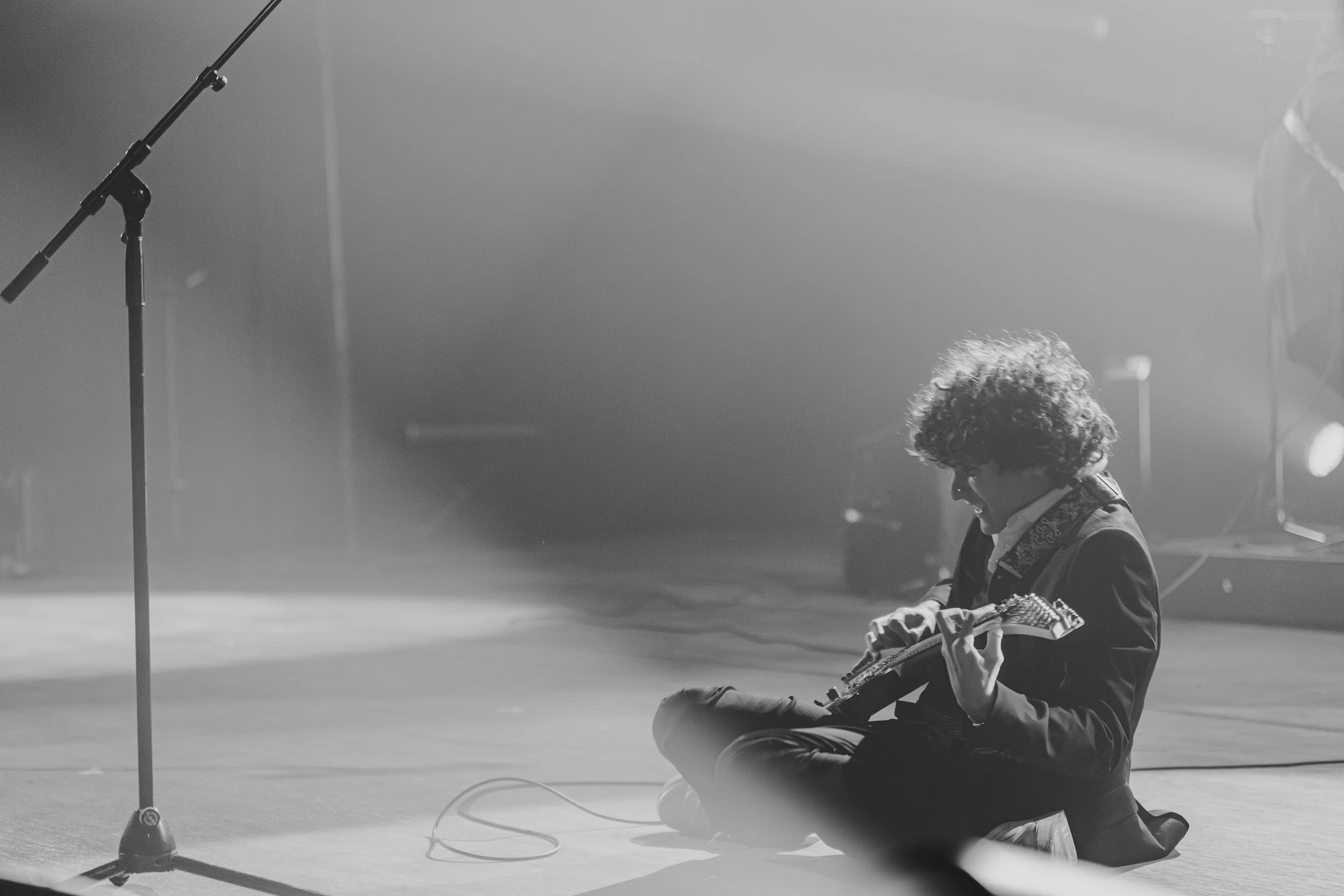 Un garçon assis par terre jouant de la guitare lors d'une performance musicale, scène sur scène avec microphone et éclairage en arrière-plan