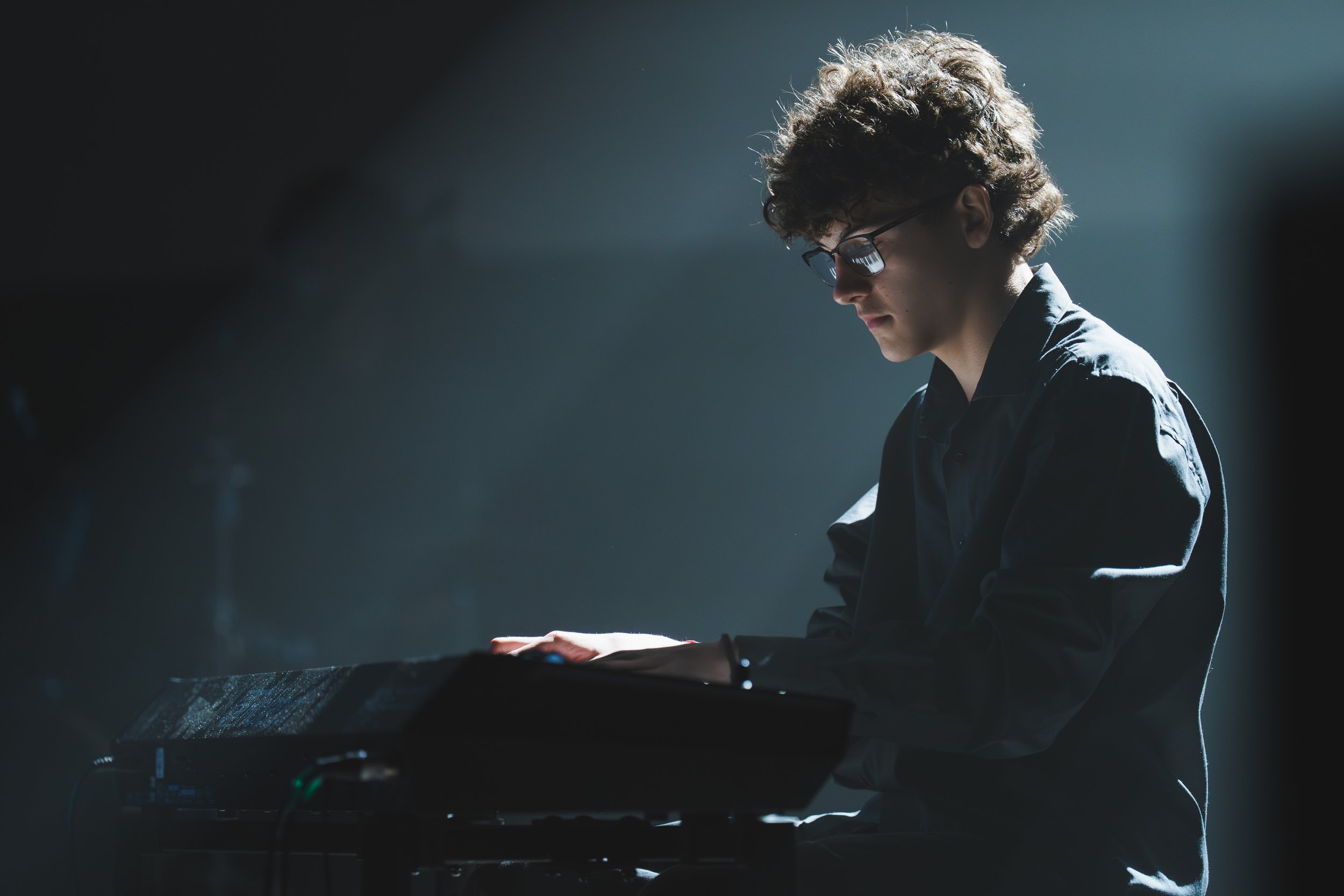 Jeune homme jouant du clavier électrique, portant des lunettes, dans un environnement sombre avec un éclairage ciblé.