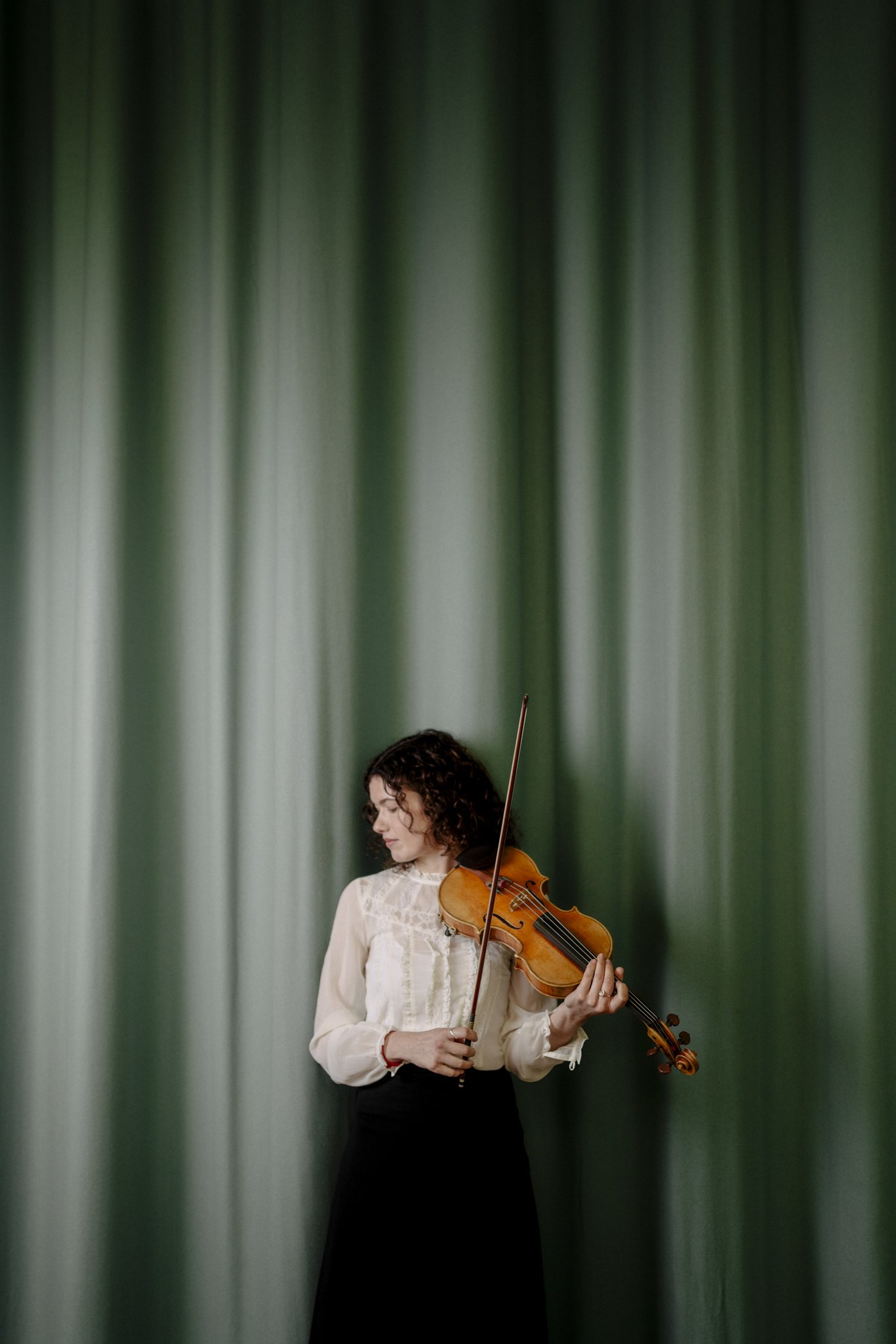 A woman standing in front of green curtains, playing a violin.