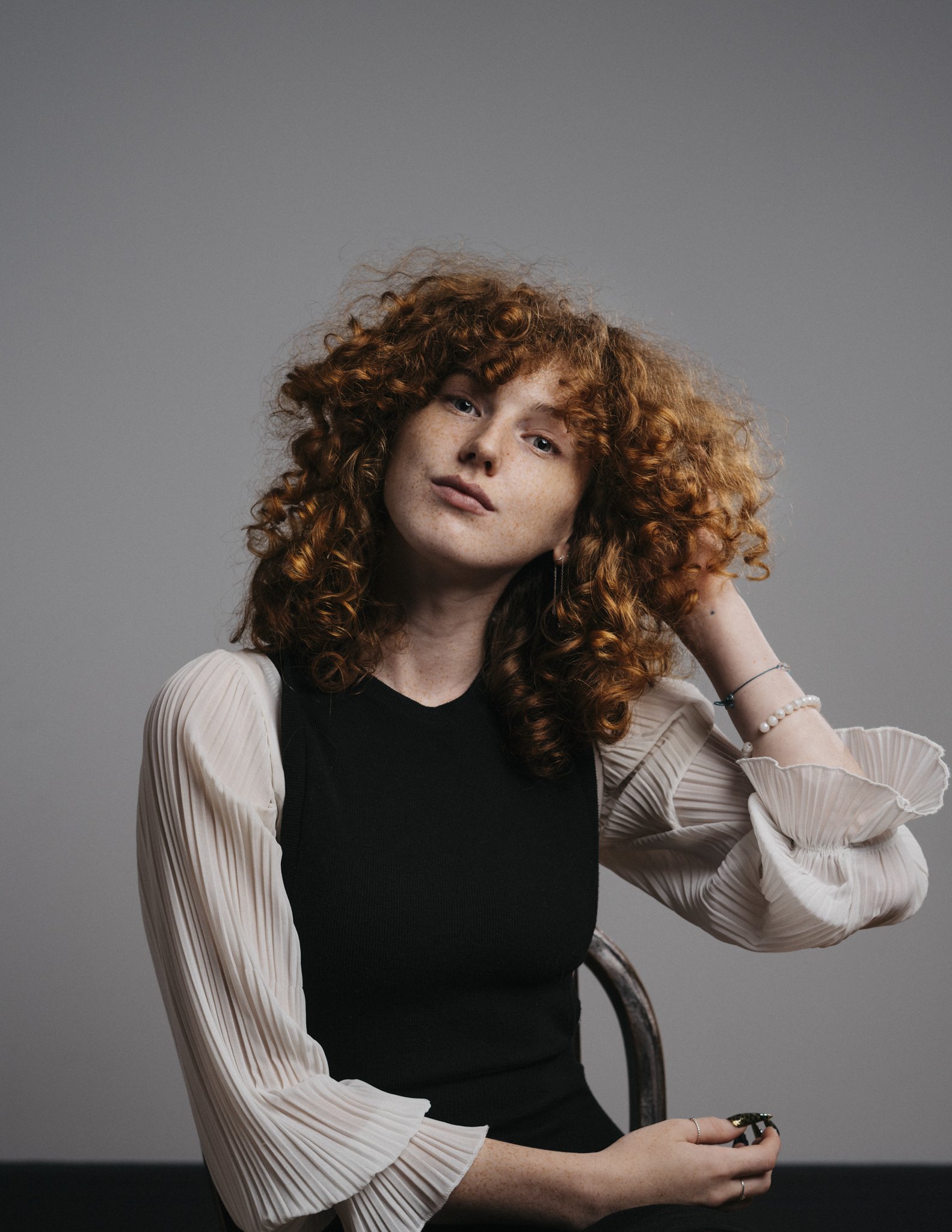 A woman with curly red hair wearing a black top and beige pleated sleeves, holding glasses and sitting on a chair, against a plain gray background.