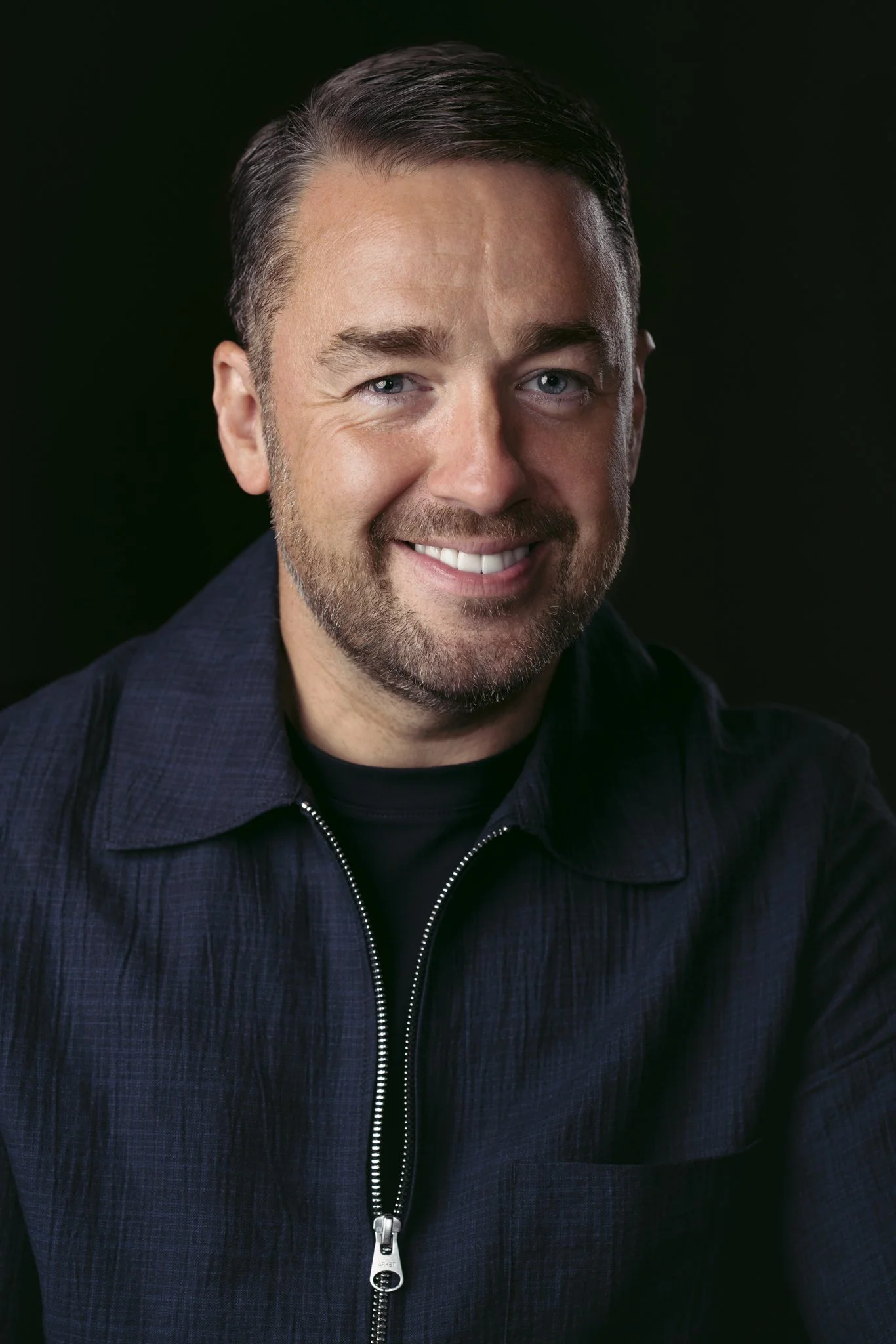 Close-up portrait of a smiling man with short, dark hair and a beard, wearing a dark zip-up jacket against a solid black background.