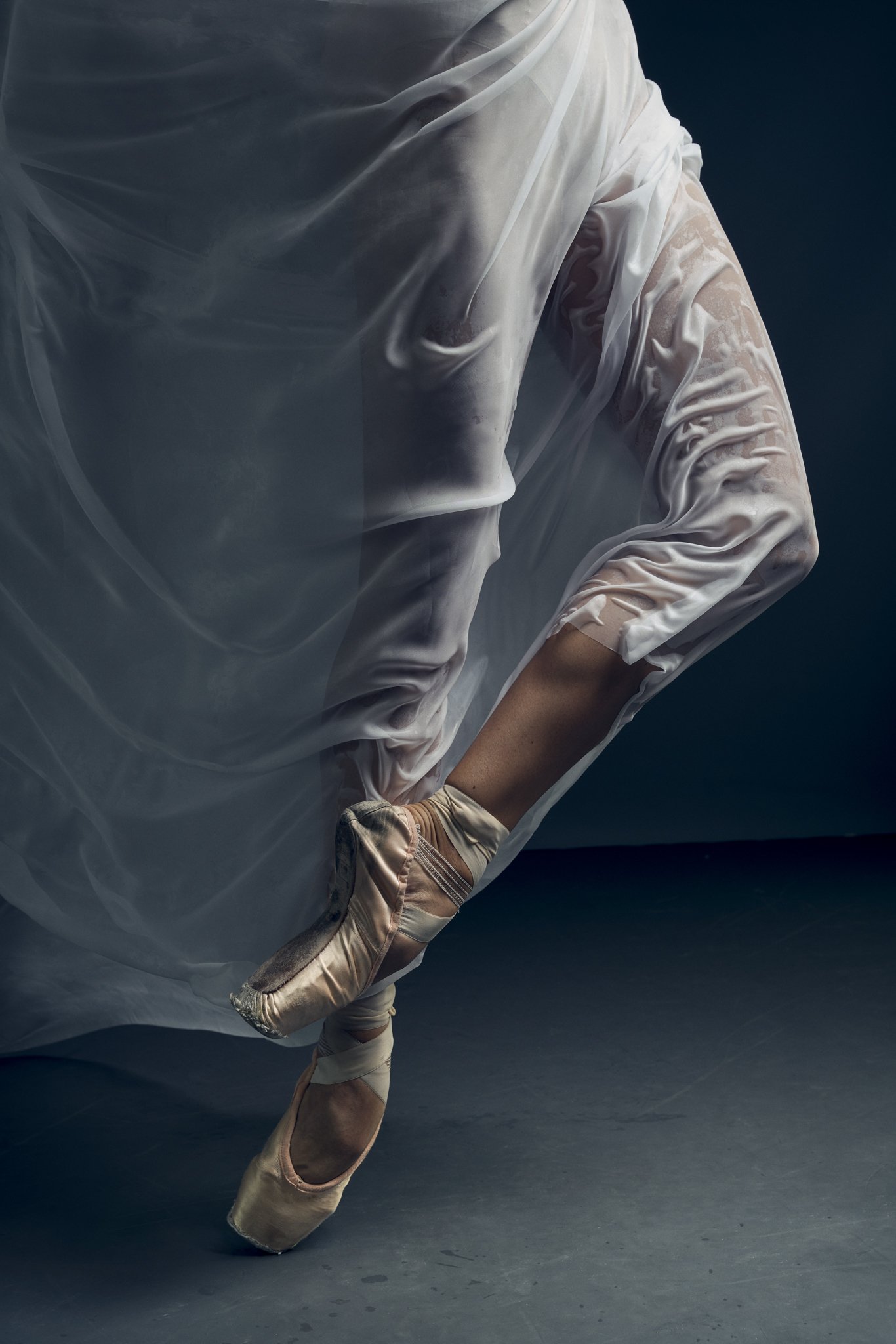 Ballet dancer in a white costume with pink pointe shoes, standing on tiptoe against a dark background.