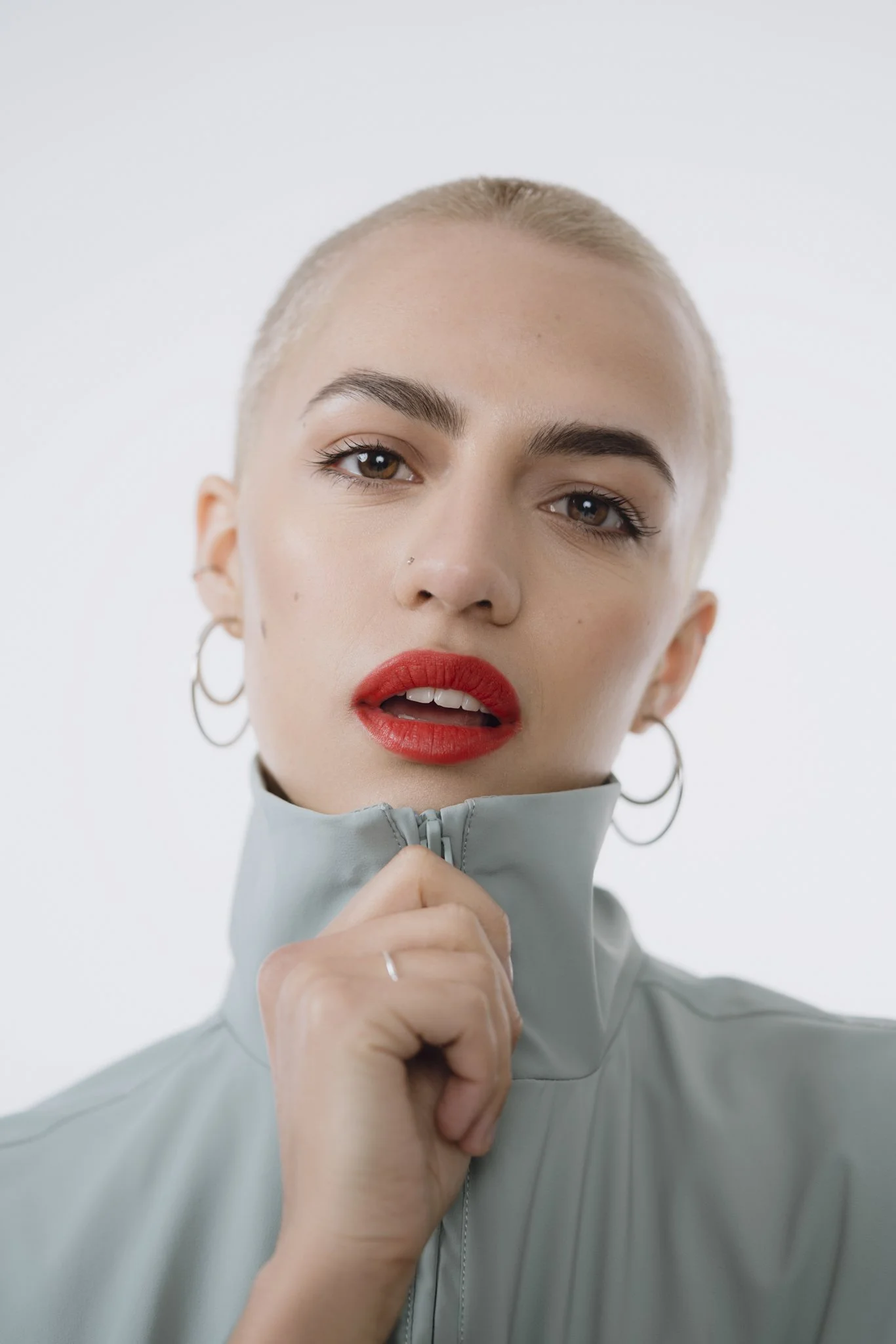 A young woman with light skin and blonde hair, tied back, is looking at the camera with a relaxed expression. She is wearing hoop earrings, red lipstick, and a light gray zip-up jacket, and her hand is holding the zipper near her chin.