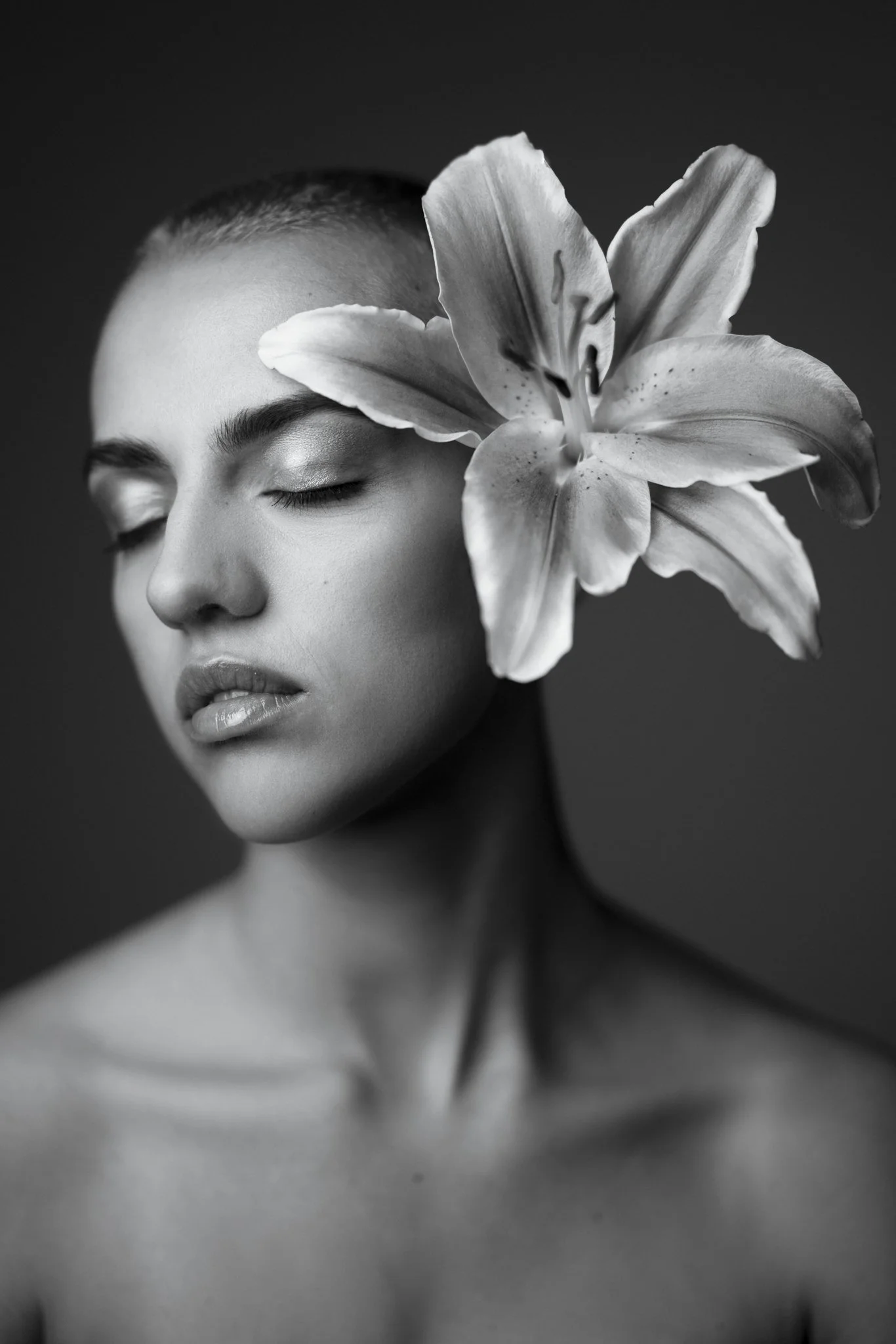 A black and white close-up portrait of a woman with closed eyes, a lily flower covering one eye and part of her face, with a dark background.