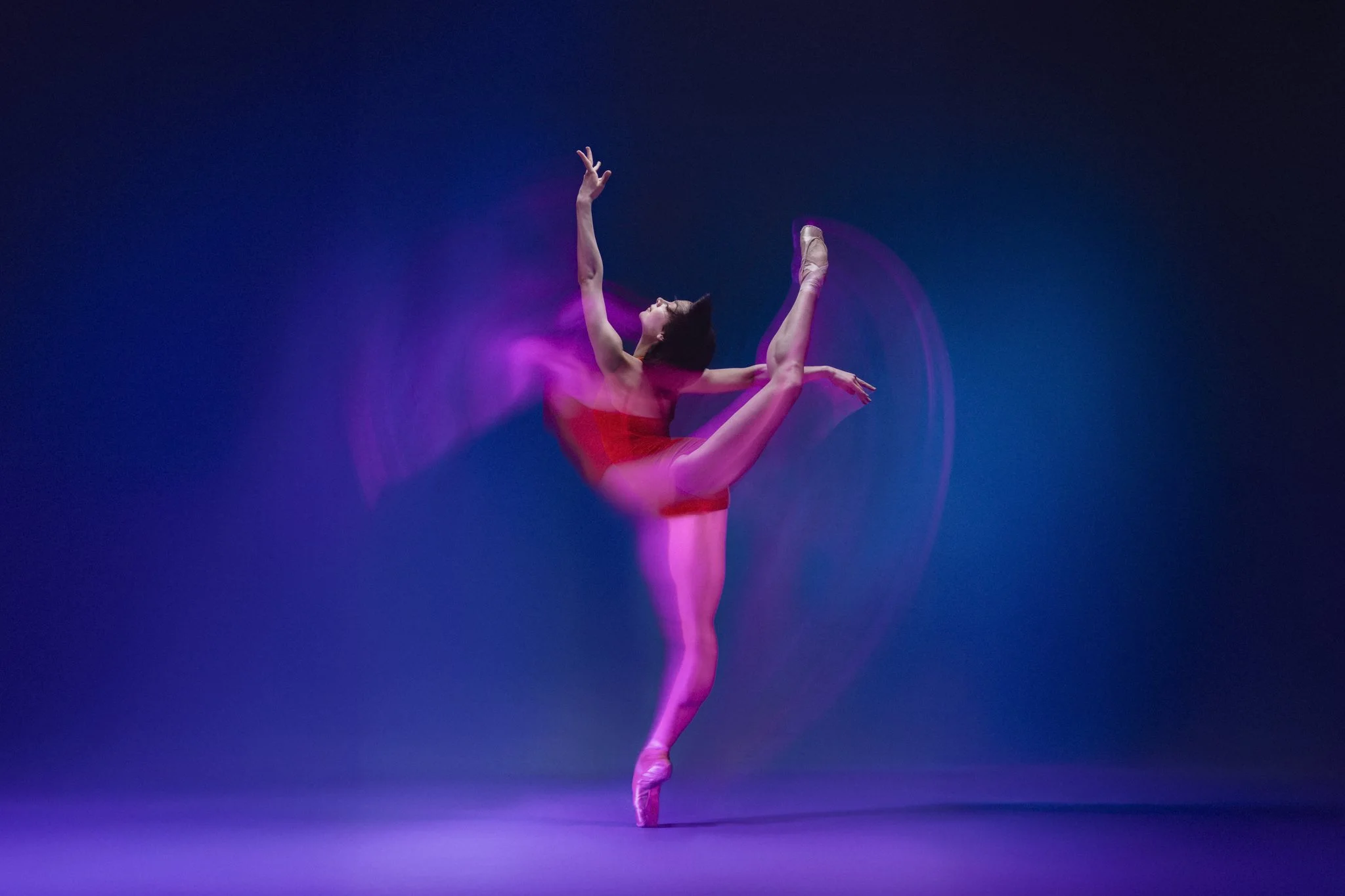 Ballet dancer in a red leotard executing a high kick on stage with blue background and colorful light effects.