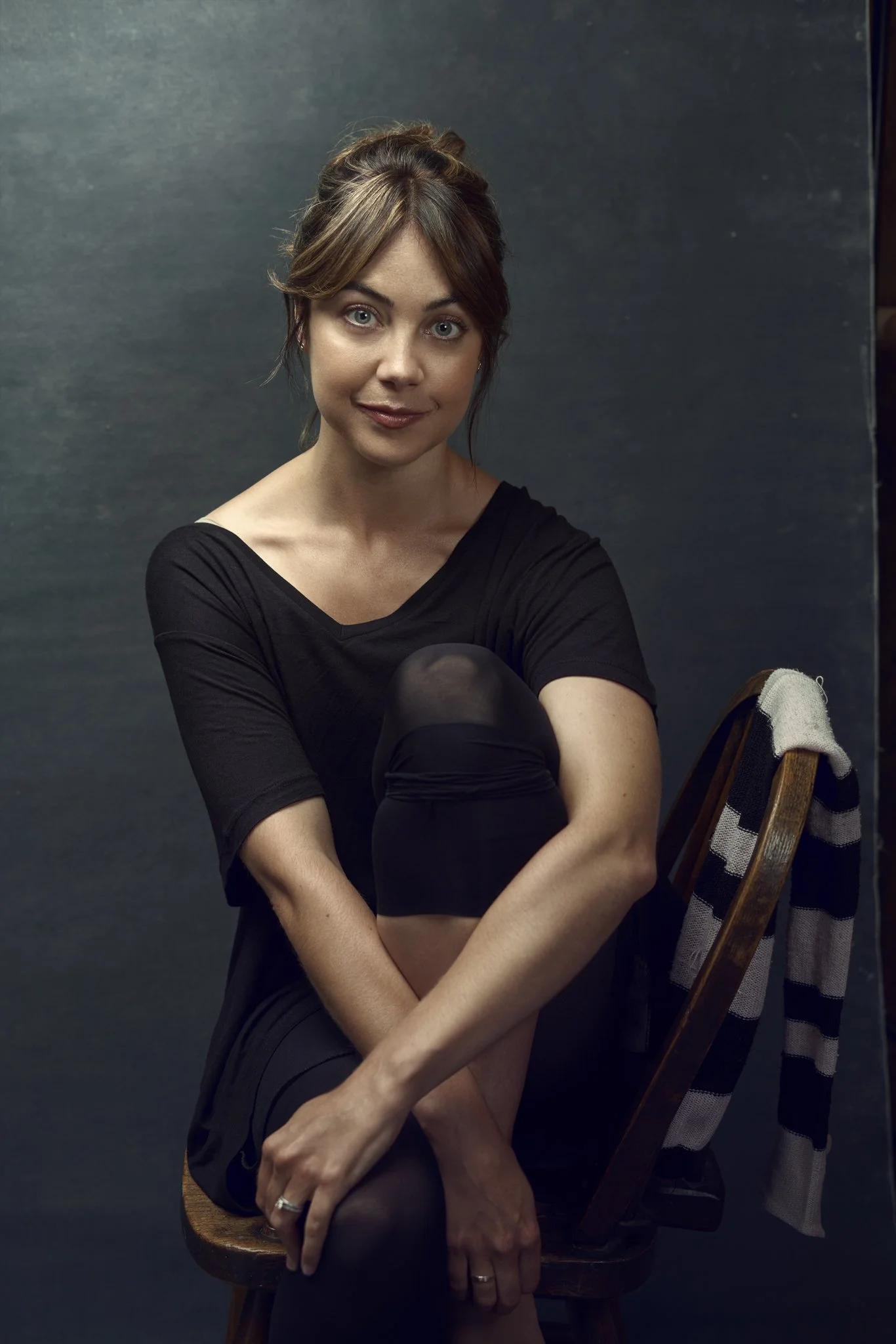 A woman sitting on a wooden chair, wearing a black dress and sheer black stockings, with her arms crossed over her knees. She has light-colored hair styled in an updo with side-swept bangs, and is smiling softly at the camera. The background is a pla