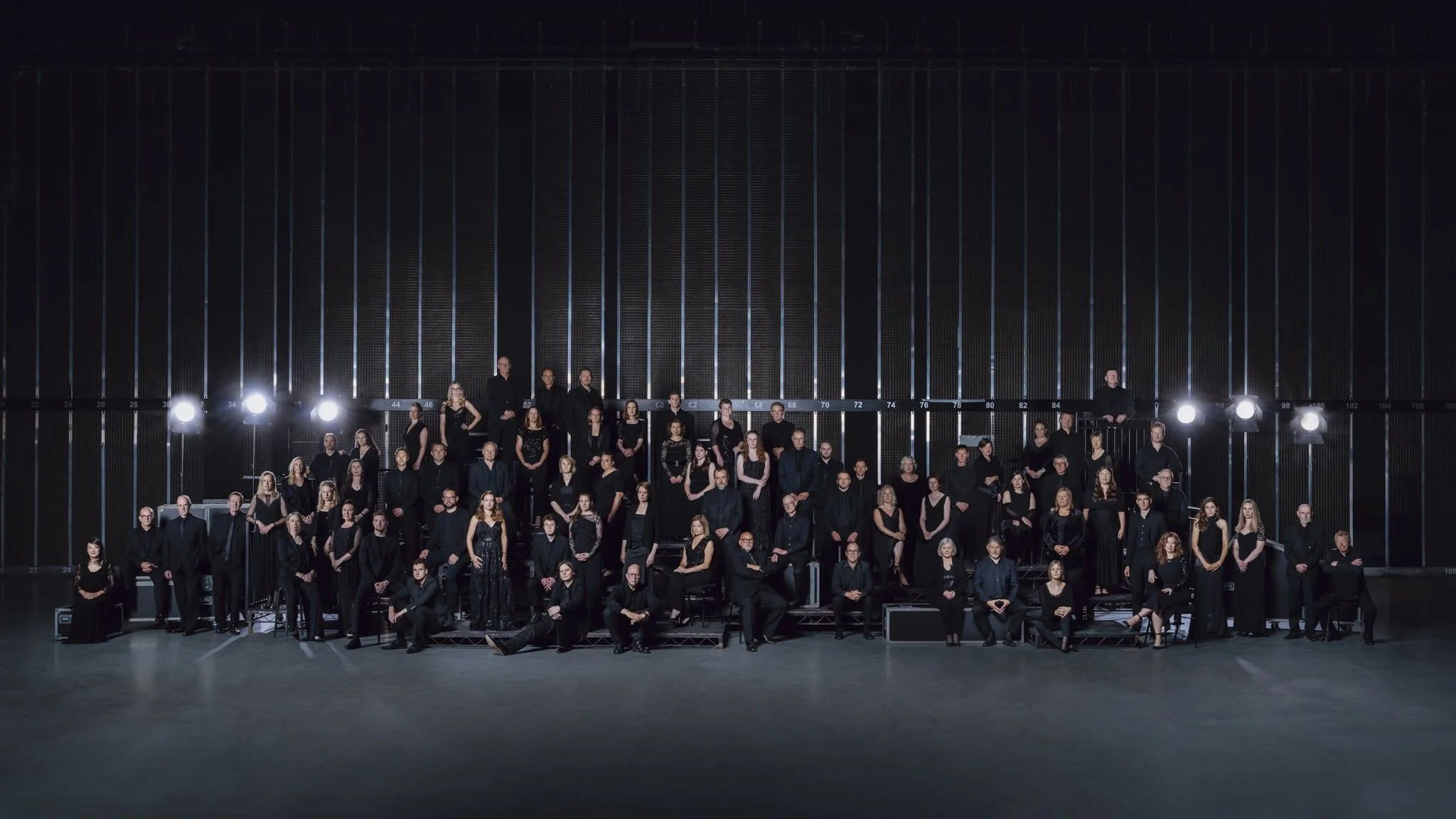 Large group portrait of people dressed in black, standing and sitting in rows inside a modern, dark architectural setting with vertical metal bars and bright lighting.