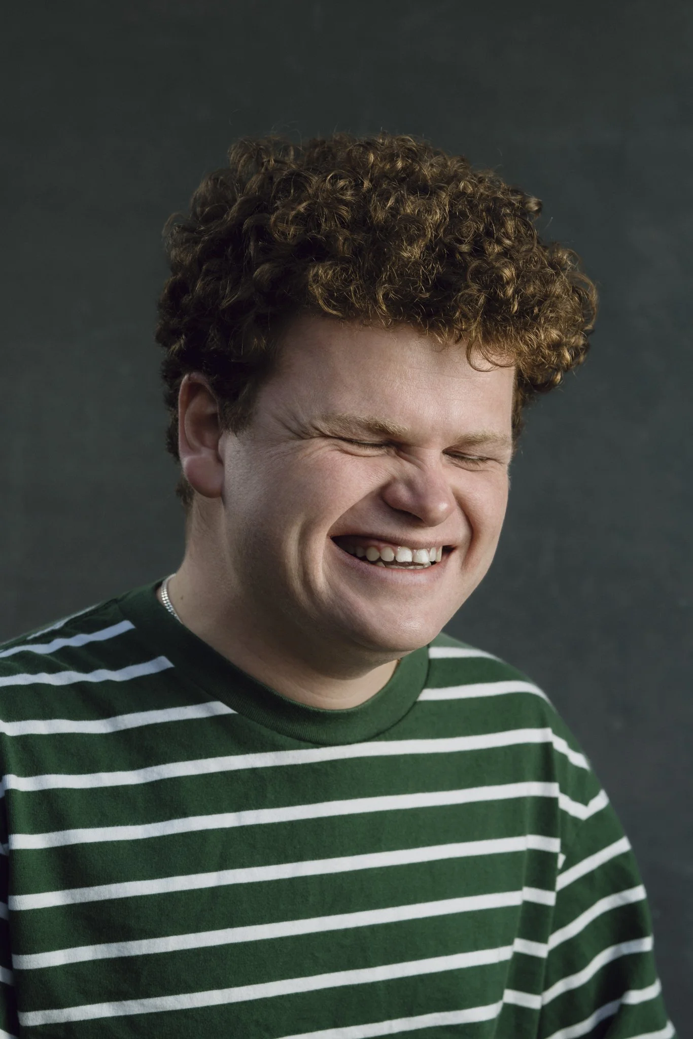 A young man with curly red hair smiling with his eyes closed, wearing a green and white striped shirt.