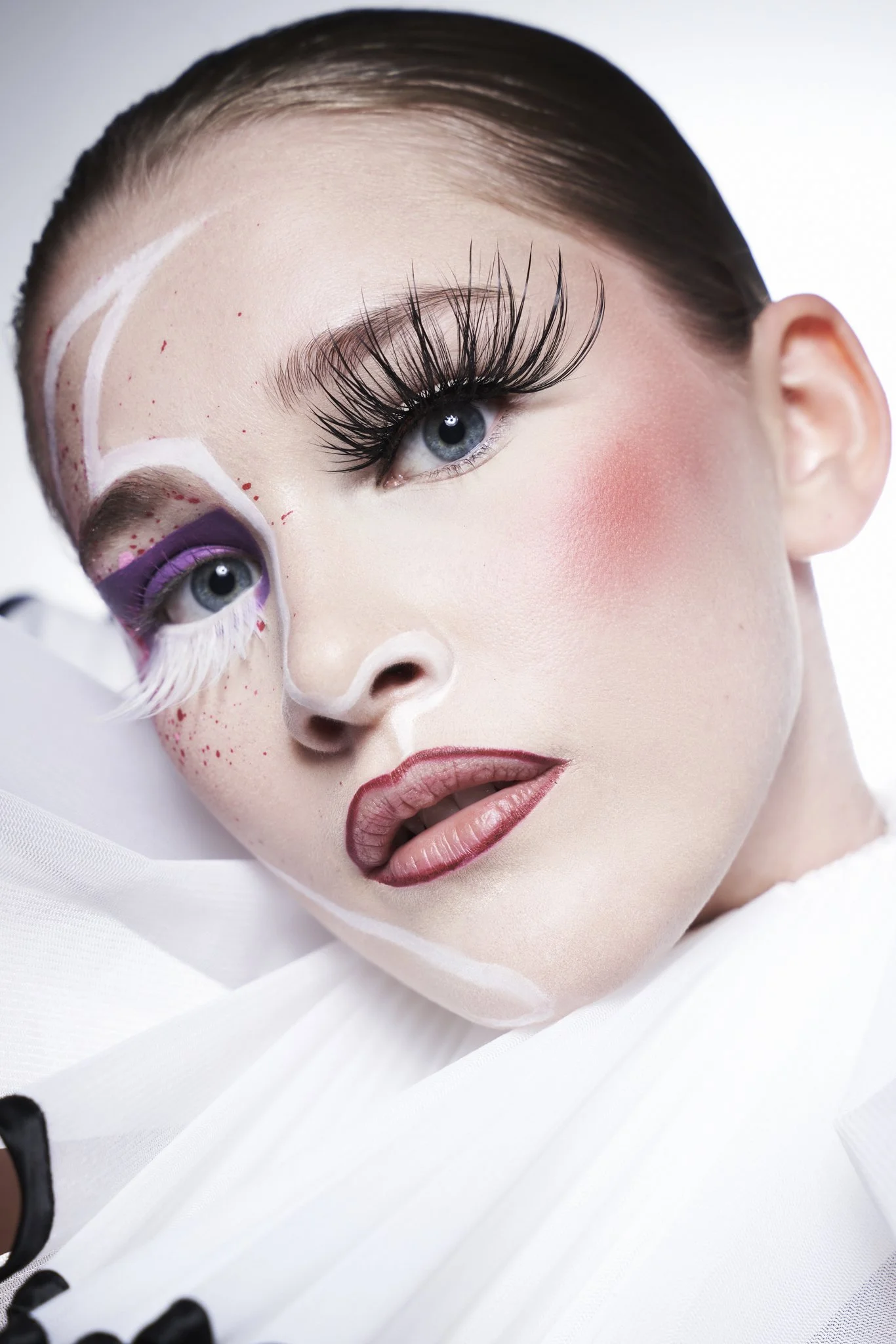 Close-up photo of a woman with elaborate makeup, including false eyelashes and pink blush, lying on a white surface with a white garment.