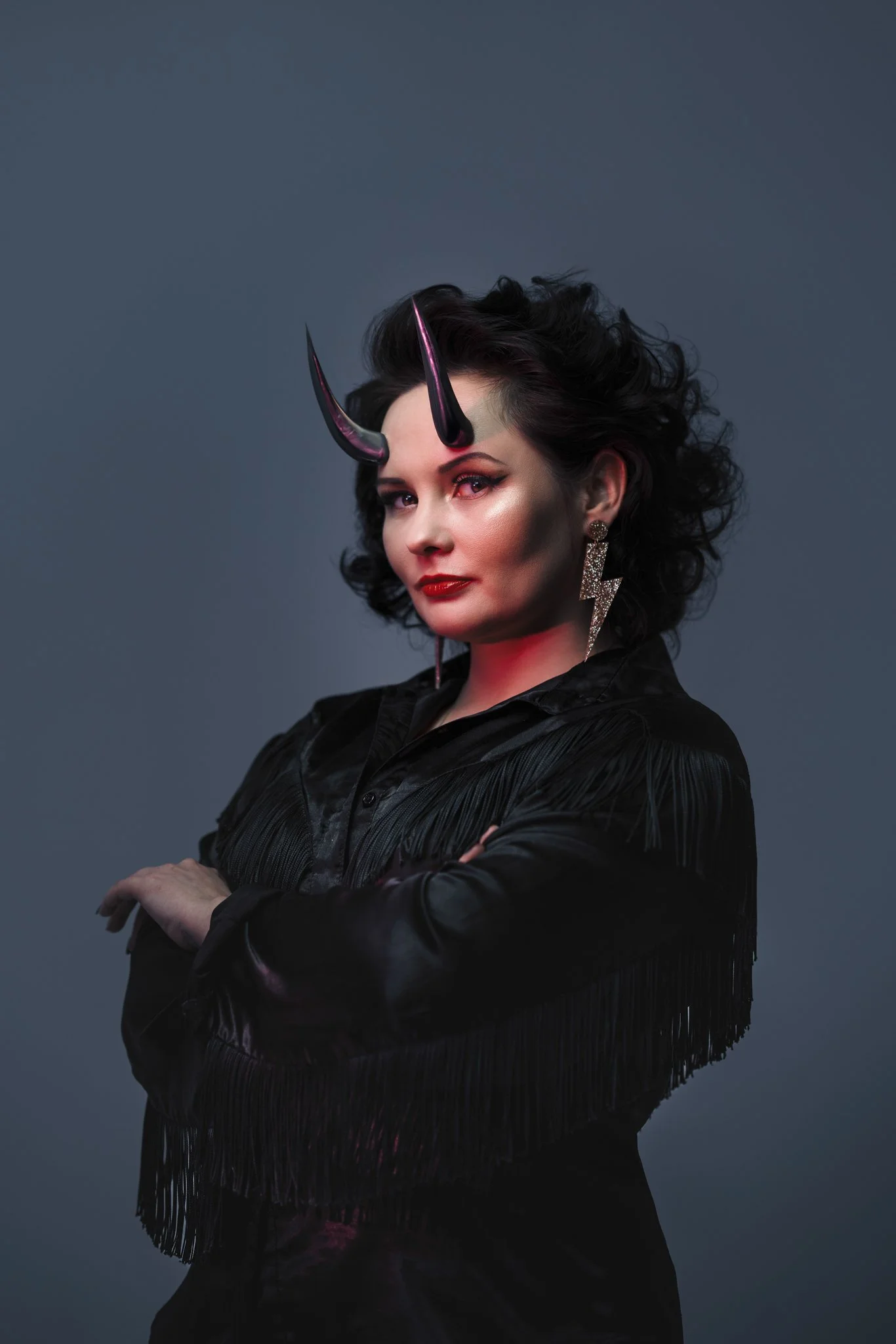A woman with dark curly hair wearing a black fringed jacket, bold red lipstick, diamond earrings in the shape of a lightning bolt, and horn-like headpieces with a pink shimmer, posing against a blue background.