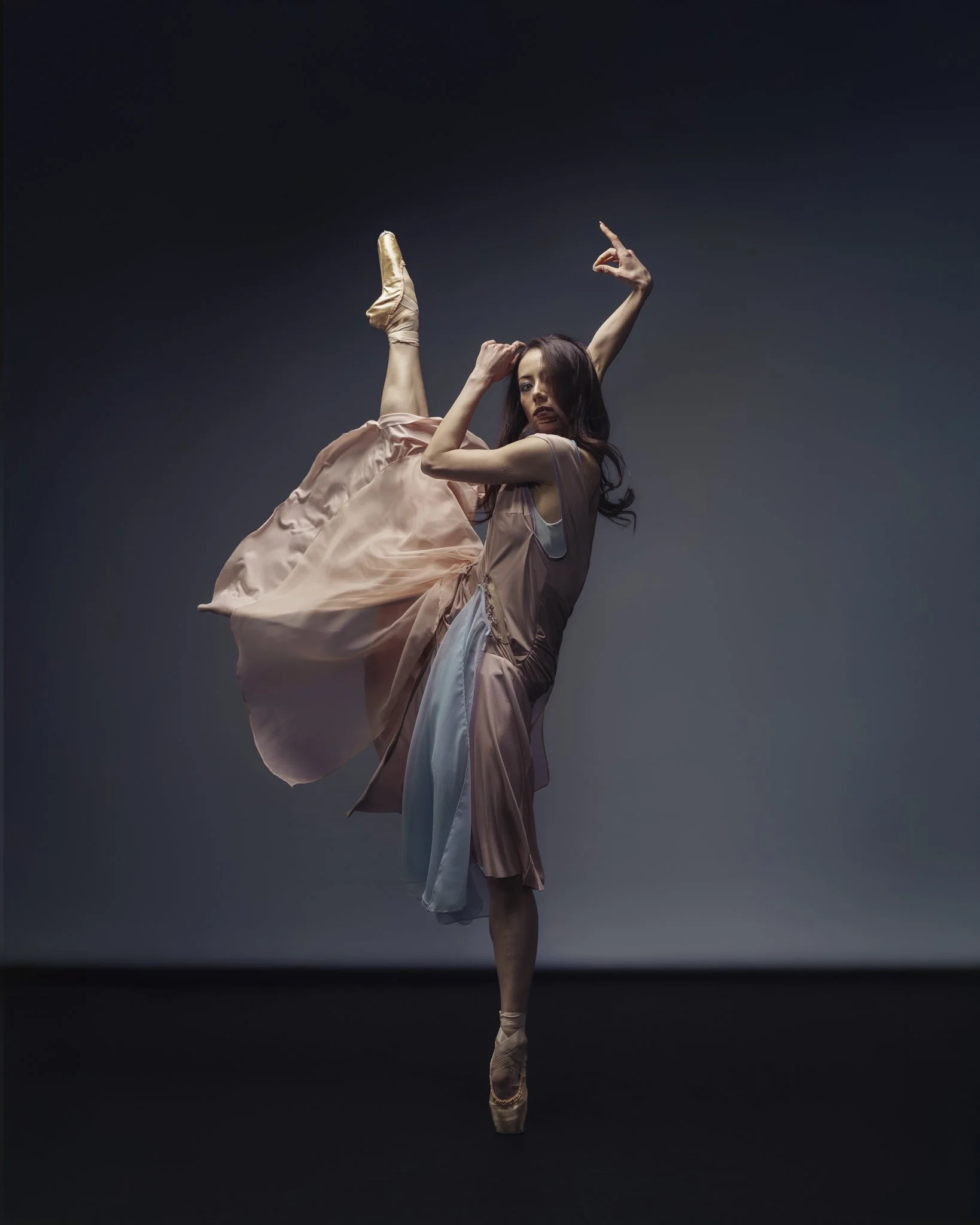 A ballet dancer performs a move on pointe against a dark background, wearing a flowing pink costume.
