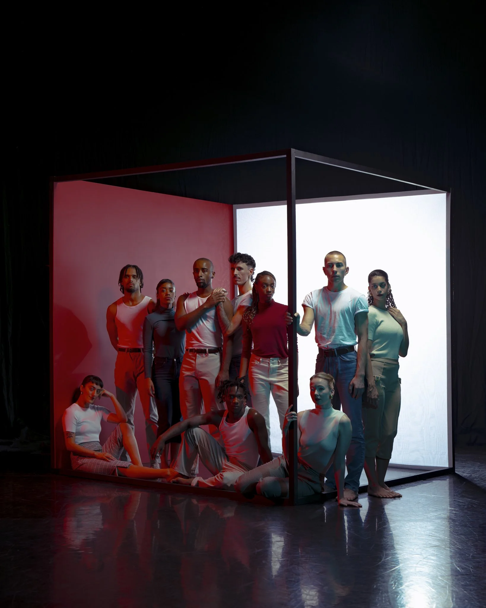 Group of diverse young people posing in front of a square art installation with black framing and illuminated white and red backgrounds, some sitting and some standing, in a contemporary art setting.