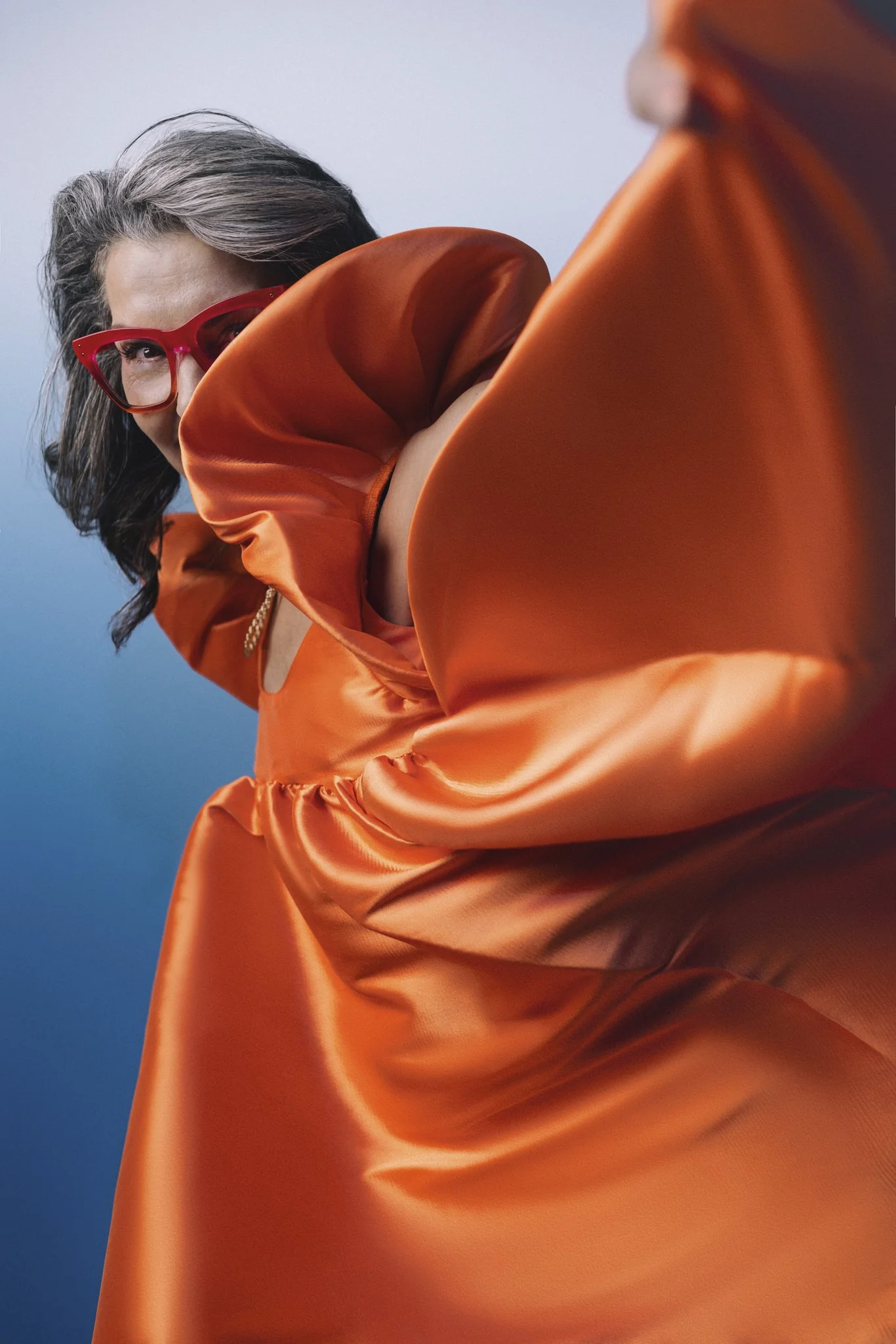 Close-up of a woman with gray hair and red glasses, wearing an orange satin dress, against a blue background, looking at the camera.