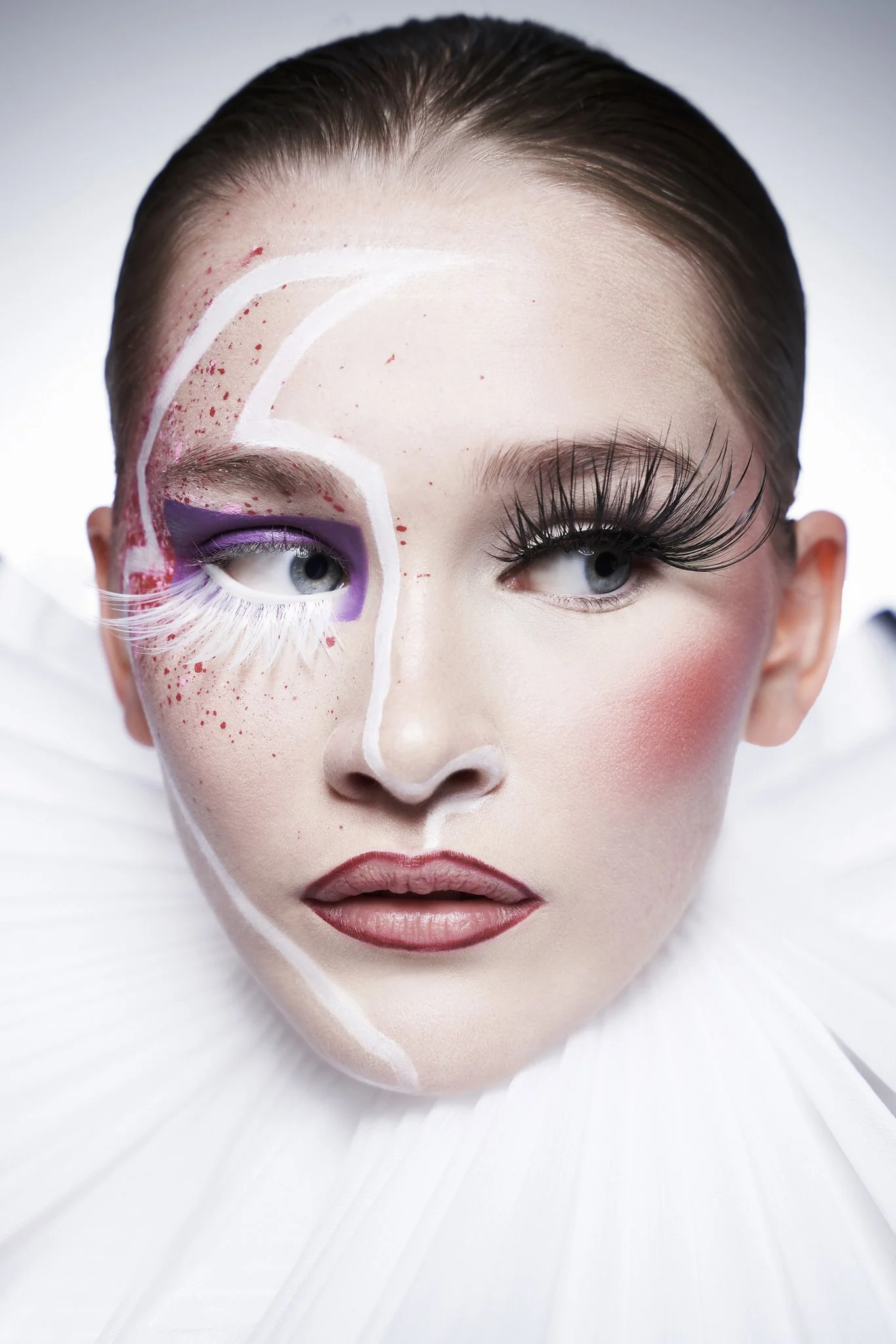Close-up of a woman's face with creative makeup and eyelash extensions, wearing a white textured fabric resembling petals or feathers.