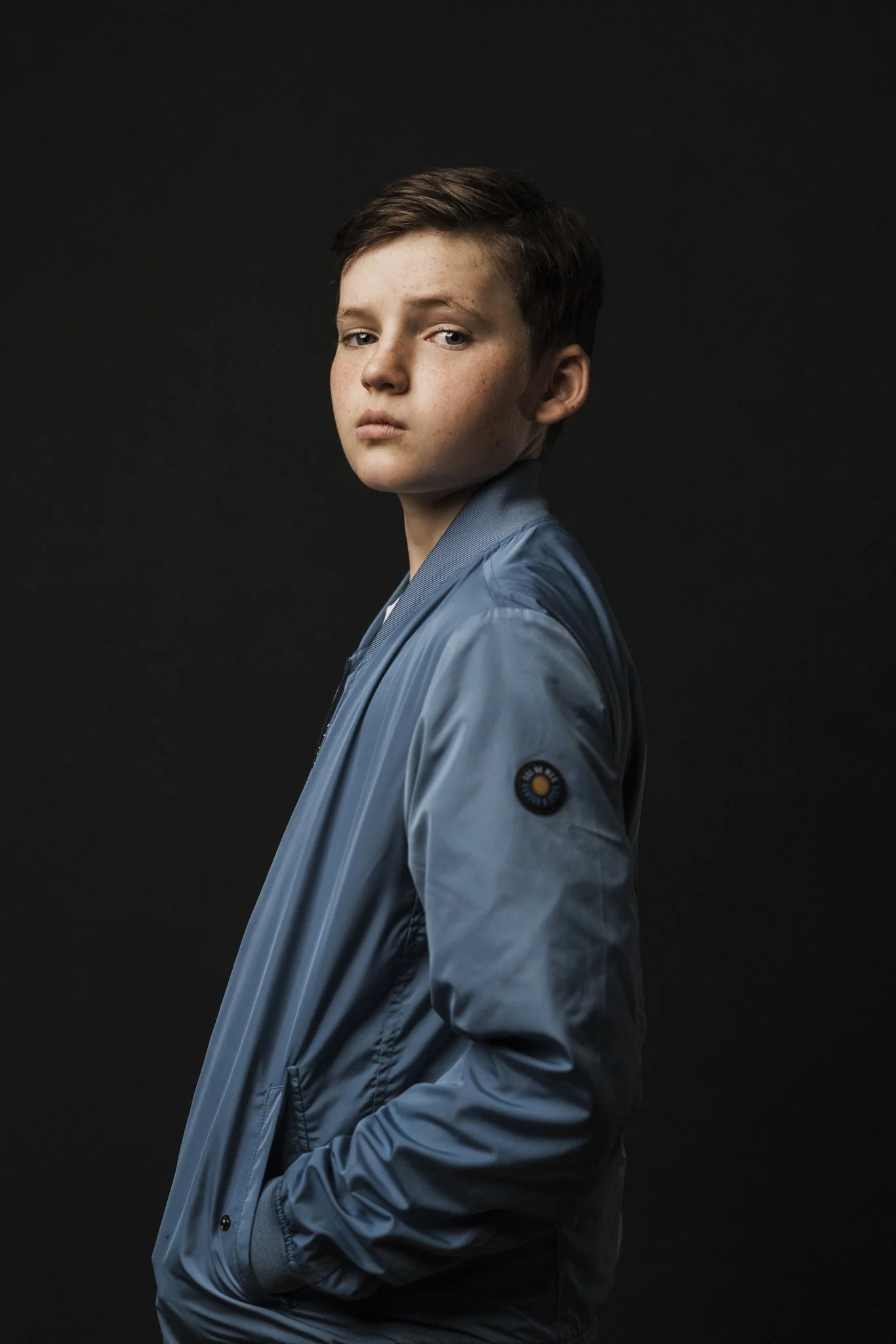 A young boy with short brown hair and fair skin wearing a light blue jacket with black buttons and a logo on the sleeve, standing against a dark background.