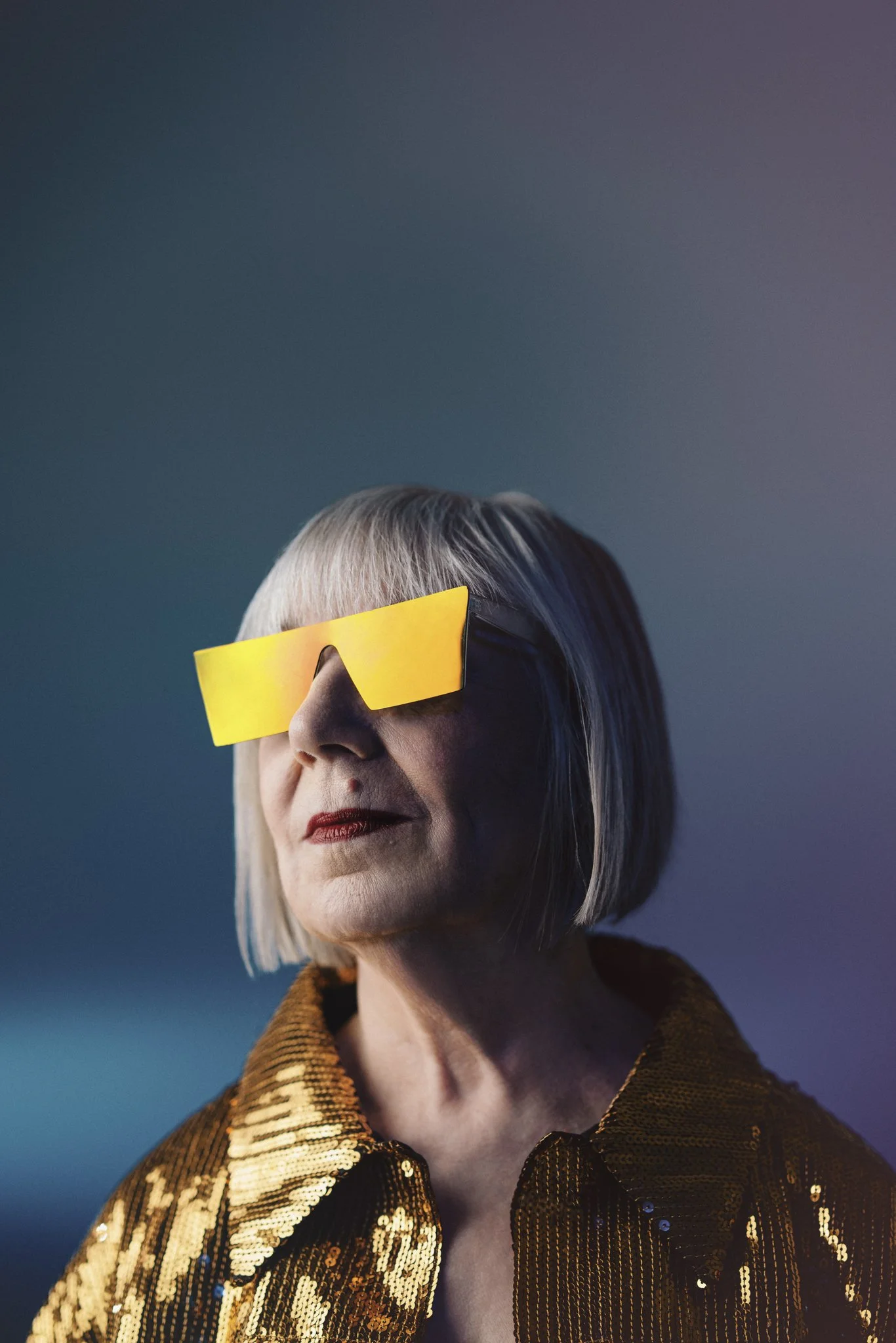 A woman with short gray hair is wearing futuristic yellow sunglasses and a shiny, gold sequin jacket against a gradient background.
