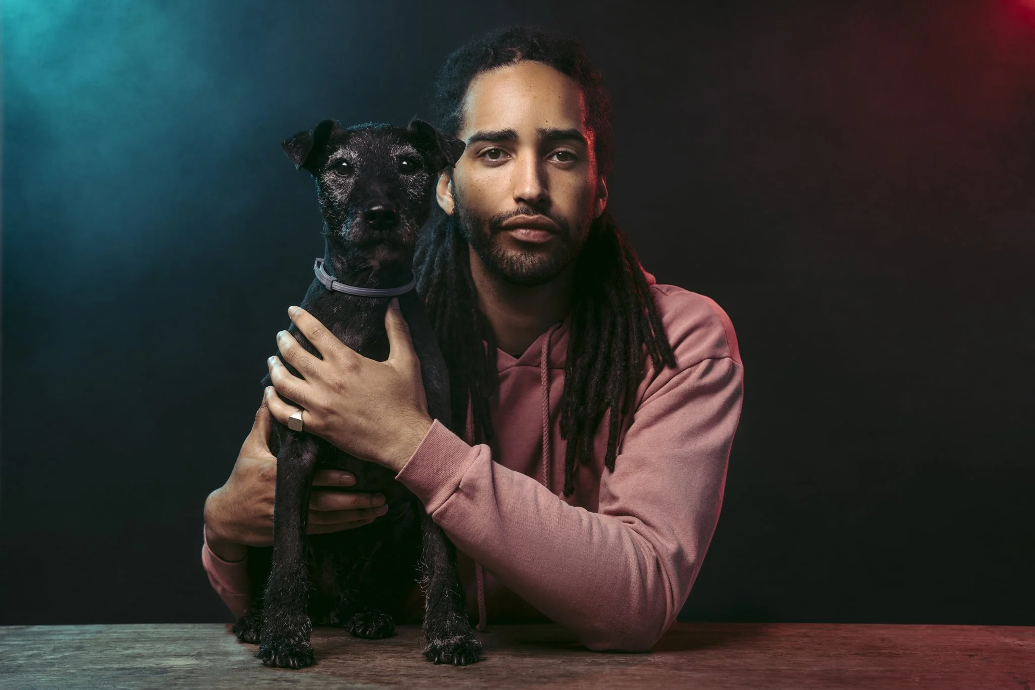A man with long dreadlocks and facial hair wearing a pink hoodie, holding a black dog with gray fur on its face, against a dark background with subtle colored lighting.