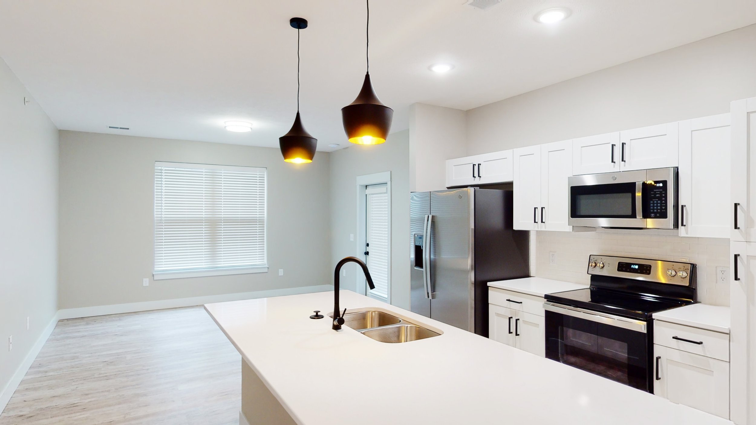 Modern kitchen and living area with white cabinets, stainless steel appliances, pendant lighting, island with sink, and large window.