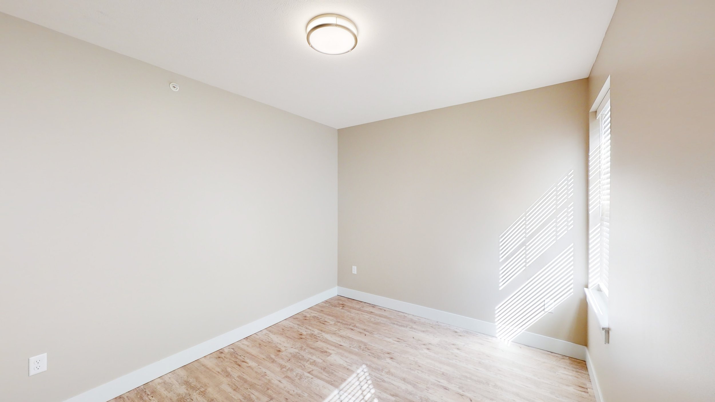 Empty room with beige walls, wooden floor, ceiling light, and window with blinds.