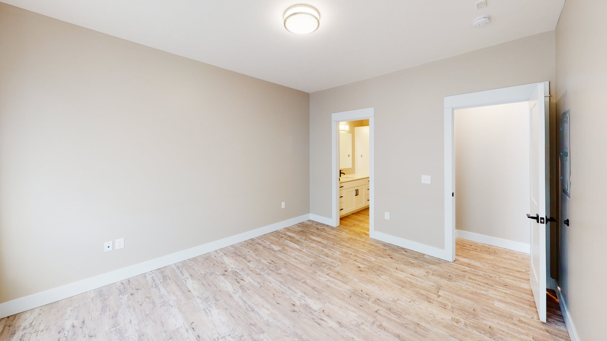Empty room with beige walls and wood flooring, featuring a ceiling light, an open door to a bathroom with a visible vanity, and another open door to an adjacent room.