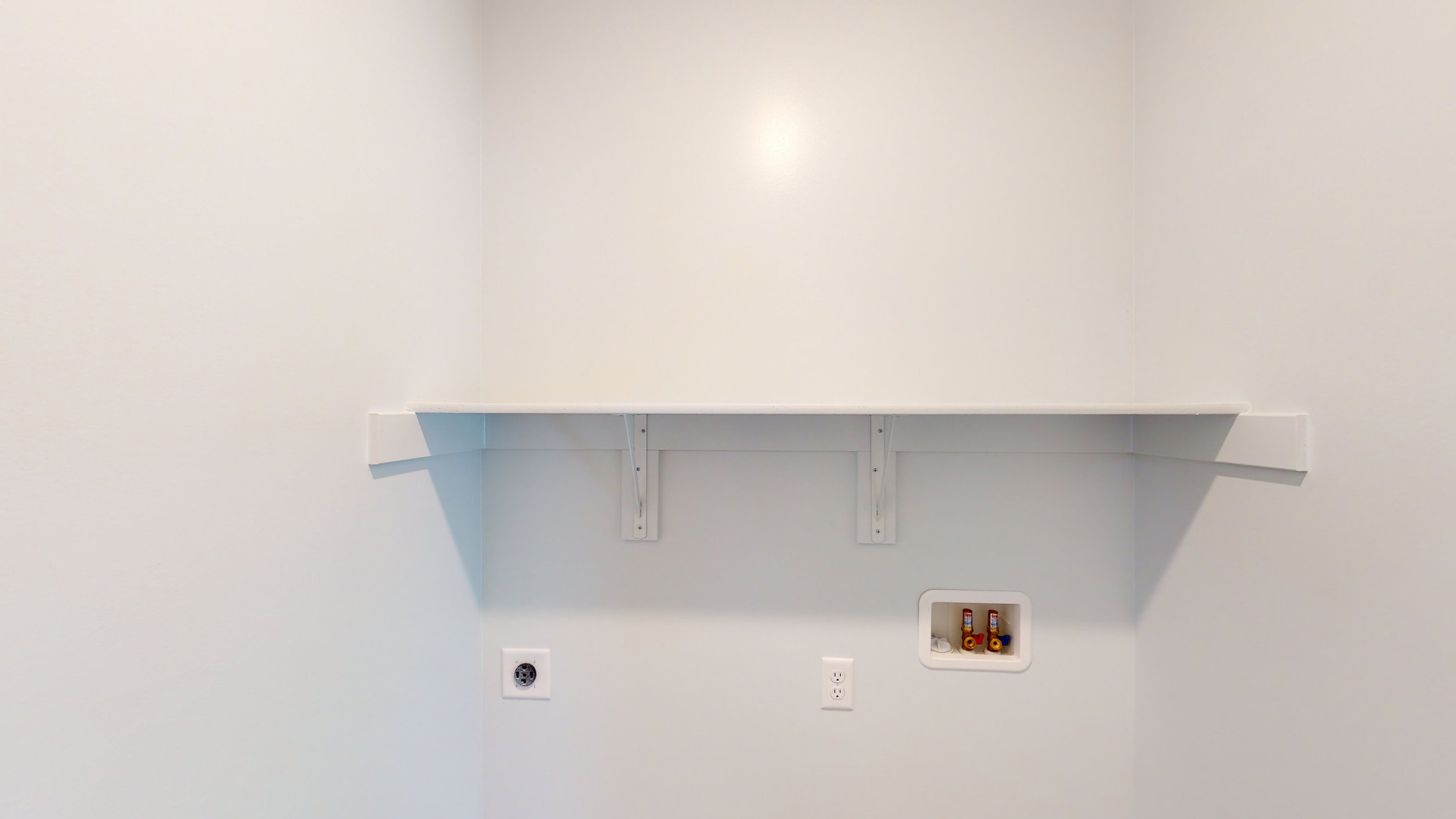 Empty white laundry room with wall shelf, electrical outlet, and water hookup