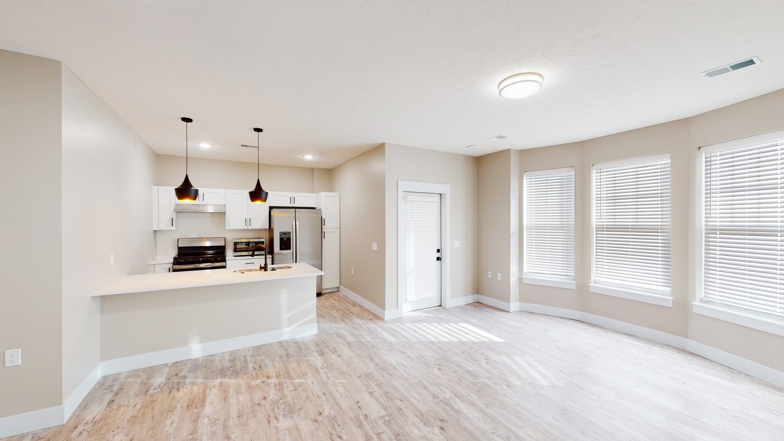 Modern unfurnished kitchen and living area with hardwood floors, white cabinets, stainless steel appliances, pendant lights, and a door leading outside. Large windows with blinds allow natural light.