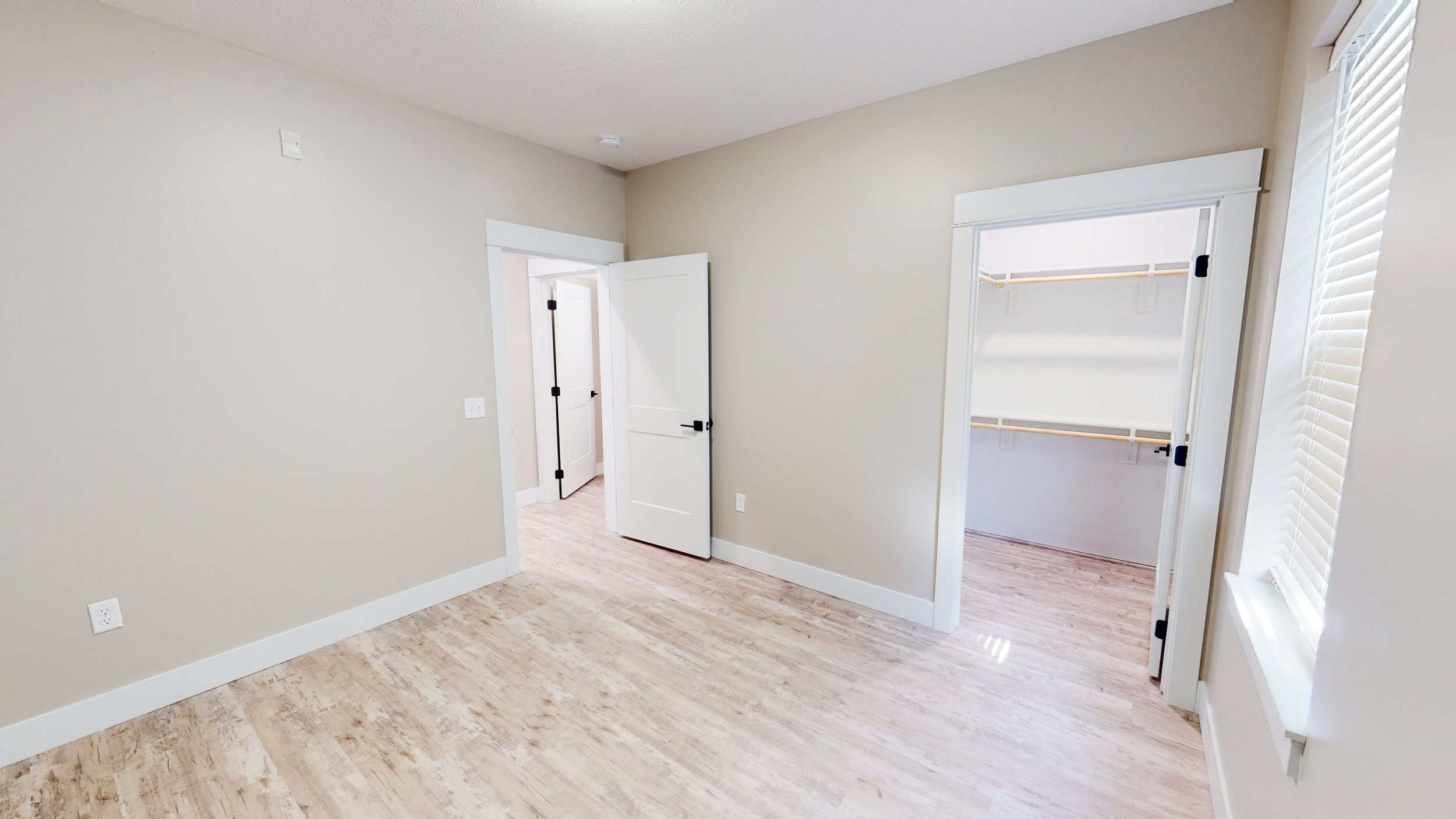 Empty room with hardwood floor, beige walls, open doorways, and a window with blinds.