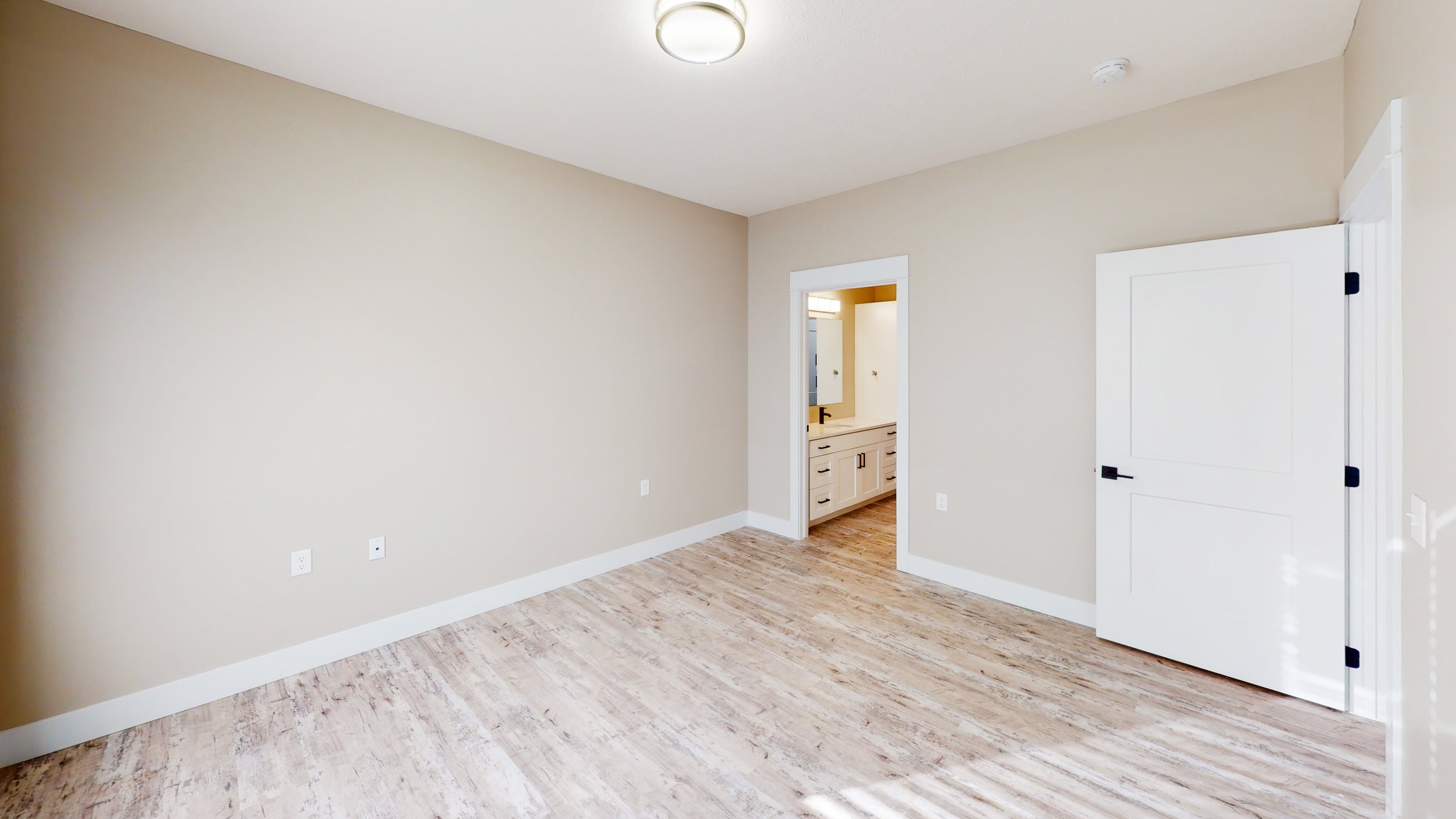 Empty room with light wood flooring, beige walls, white door, and a view into a bathroom with white cabinets.