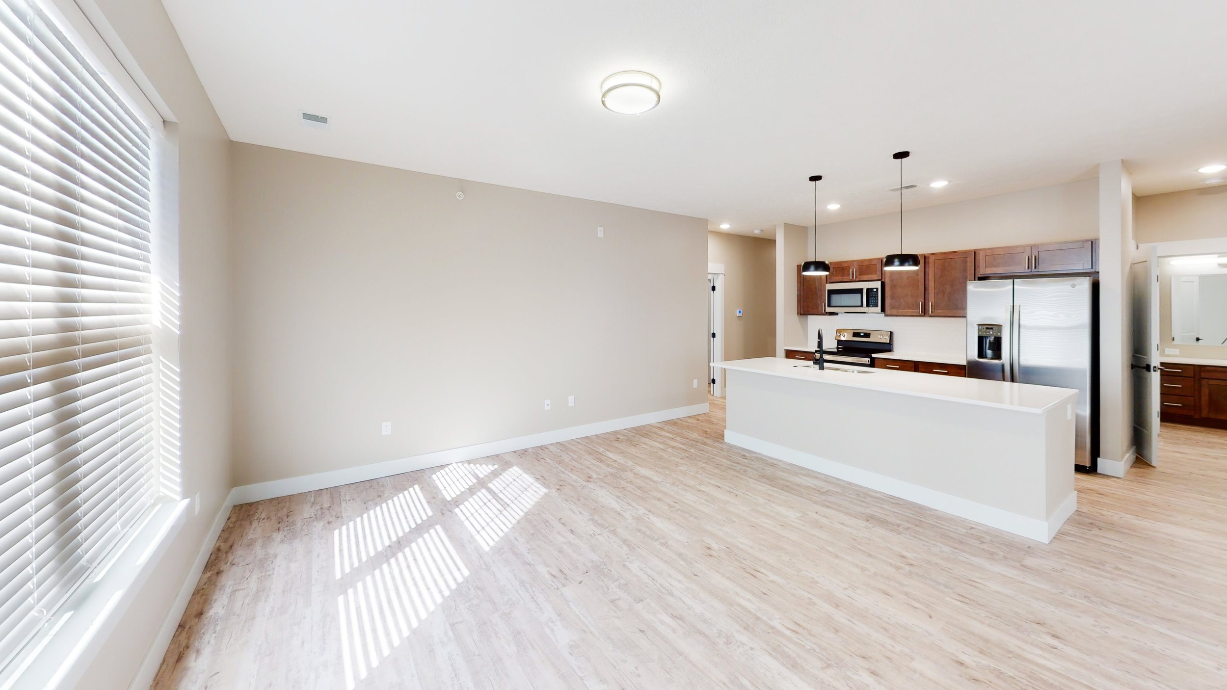 Modern open-plan kitchen and living area with wooden flooring, white walls, and large window with blinds. Features include stainless steel appliances, wooden cabinets, and pendant lights over the kitchen island.