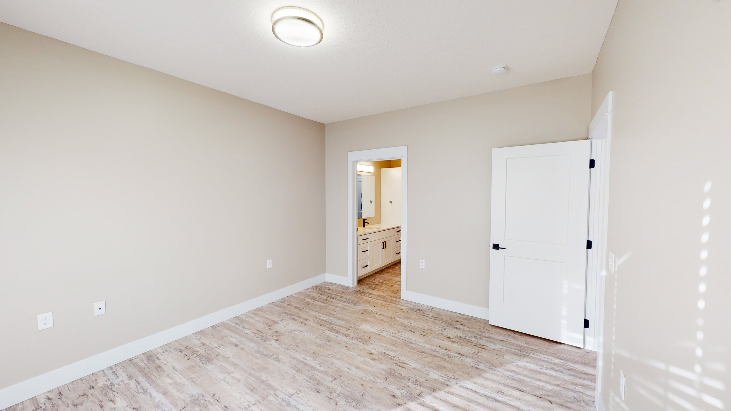 Empty room with beige walls, light wood flooring, ceiling light fixture, and a doorway leading to a bathroom with white cabinets.