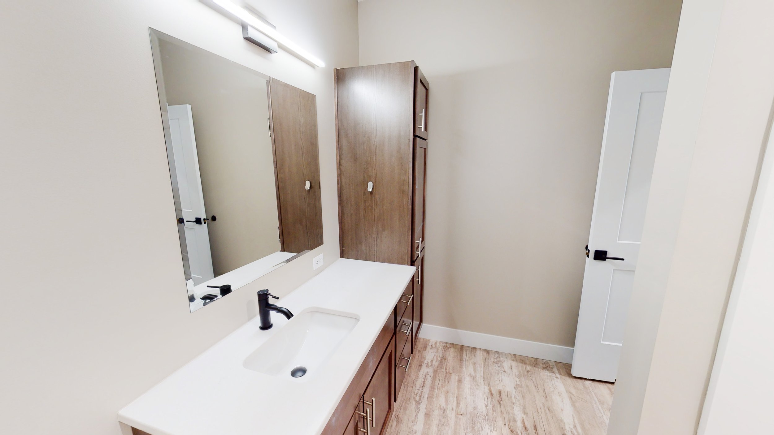 Modern bathroom with a large mirror, wooden cabinet, white countertop, sink with black faucet, and an open door.
