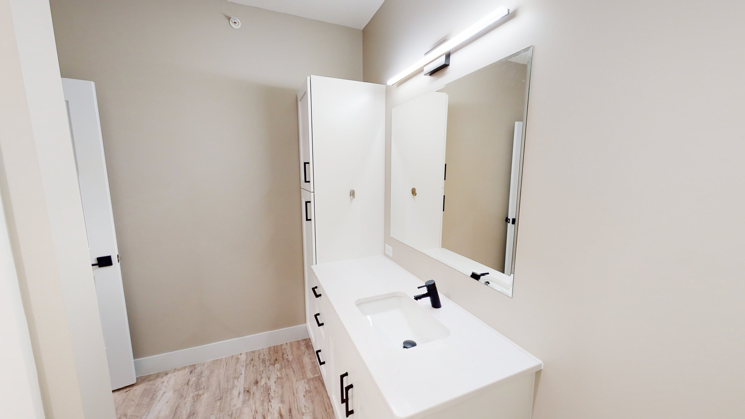 Modern bathroom with white vanity, large mirror, black fixtures, and beige walls.