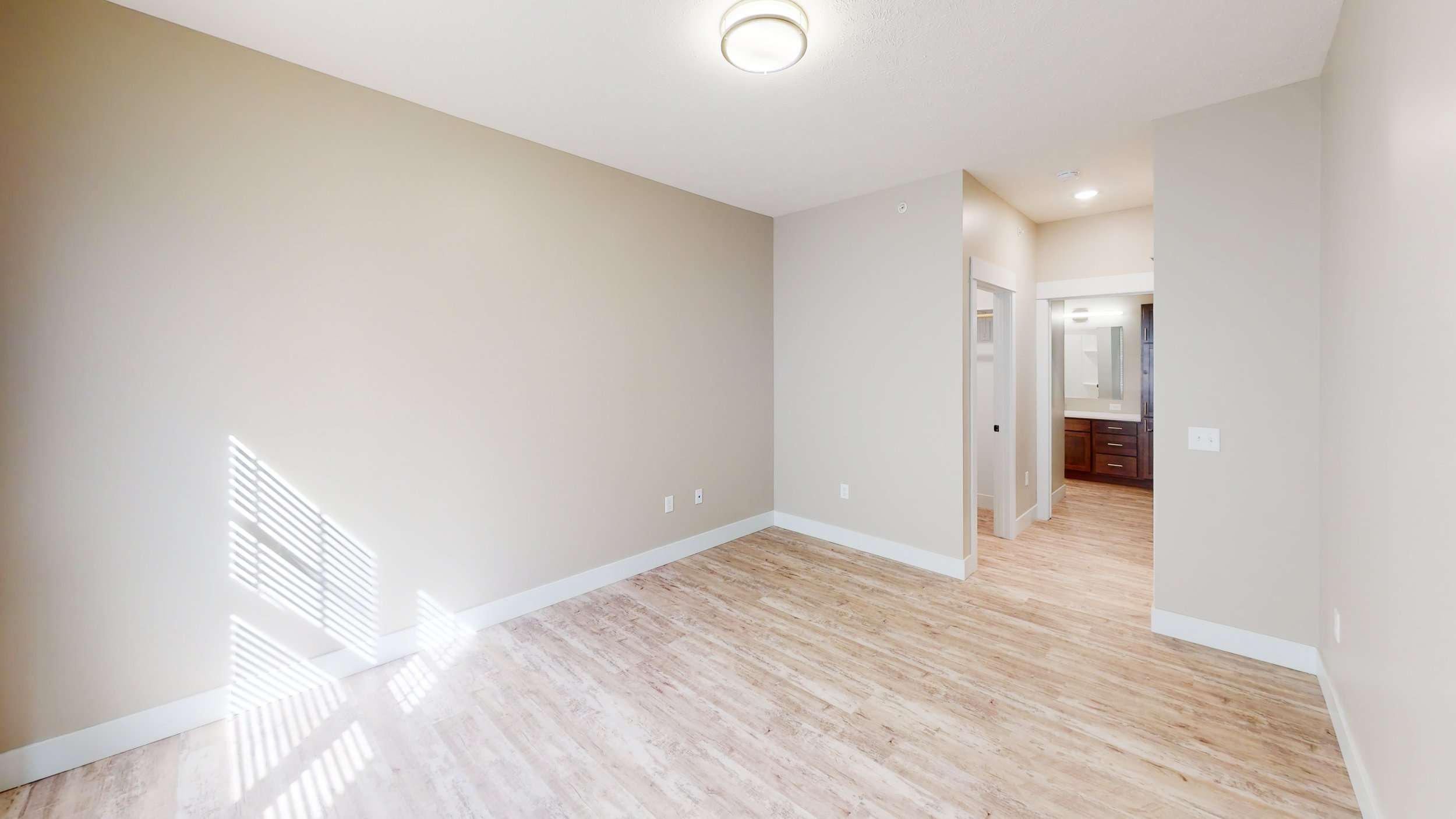 Empty room with light wood flooring, beige walls, and ceiling light, connected to a bathroom with a sink and cabinets.
